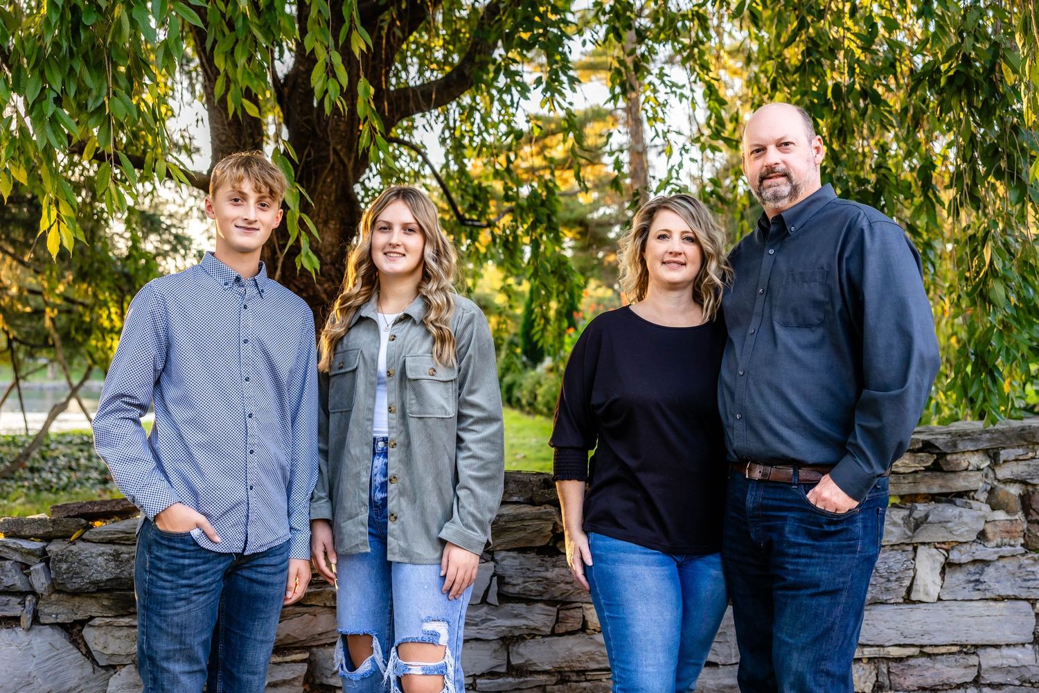 Family of four poses together against stone wall and willow trees during golden hour.