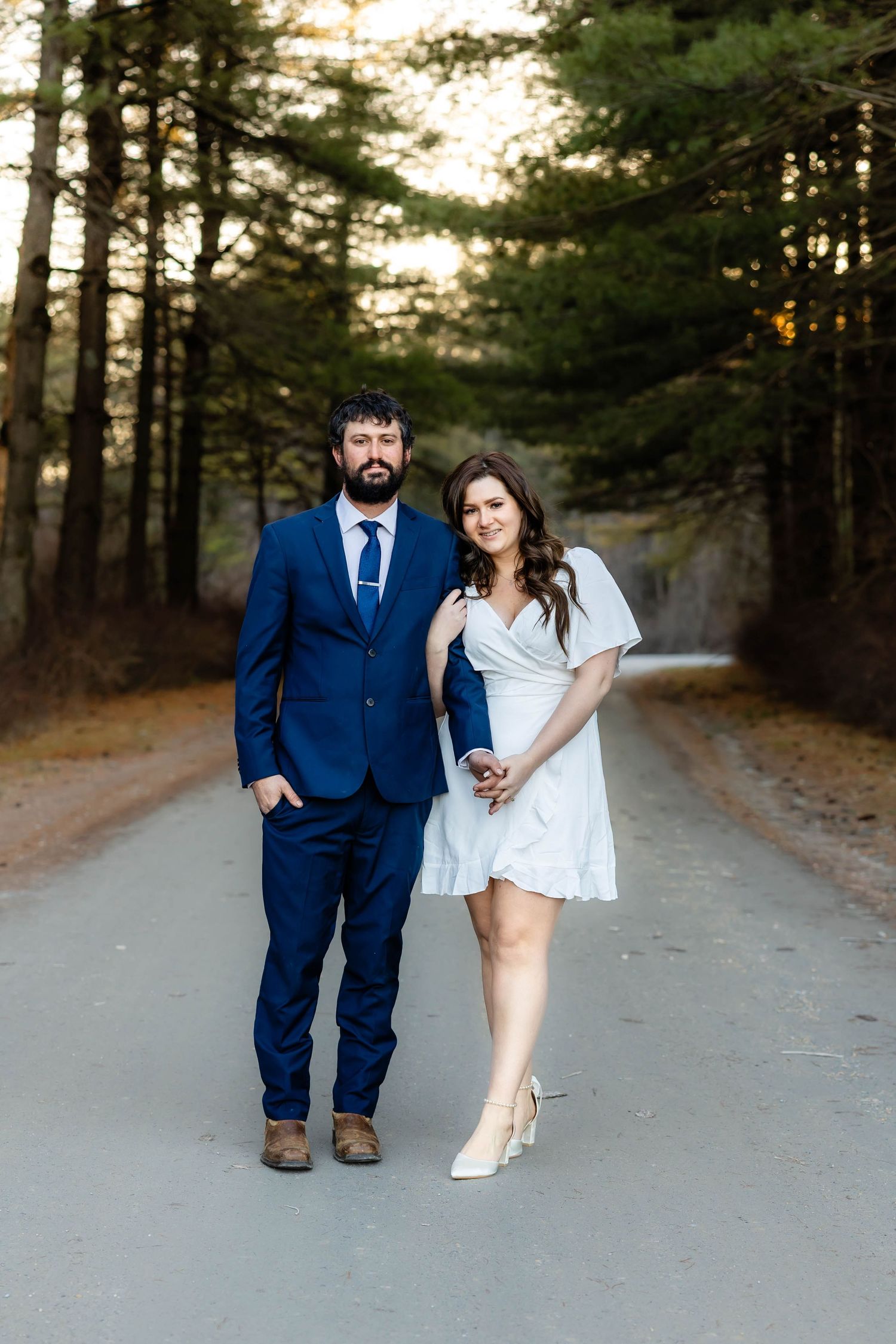 A couple stands together on a tree-lined path at sunset, wearing a blue suit and white dress.