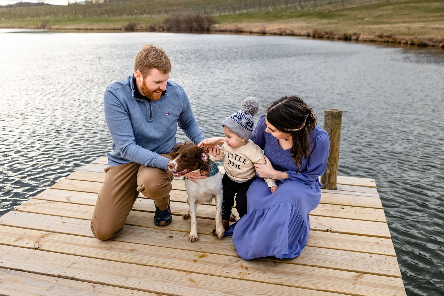 A family plays with their small dog on a wooden dock overlooking a serene lake.