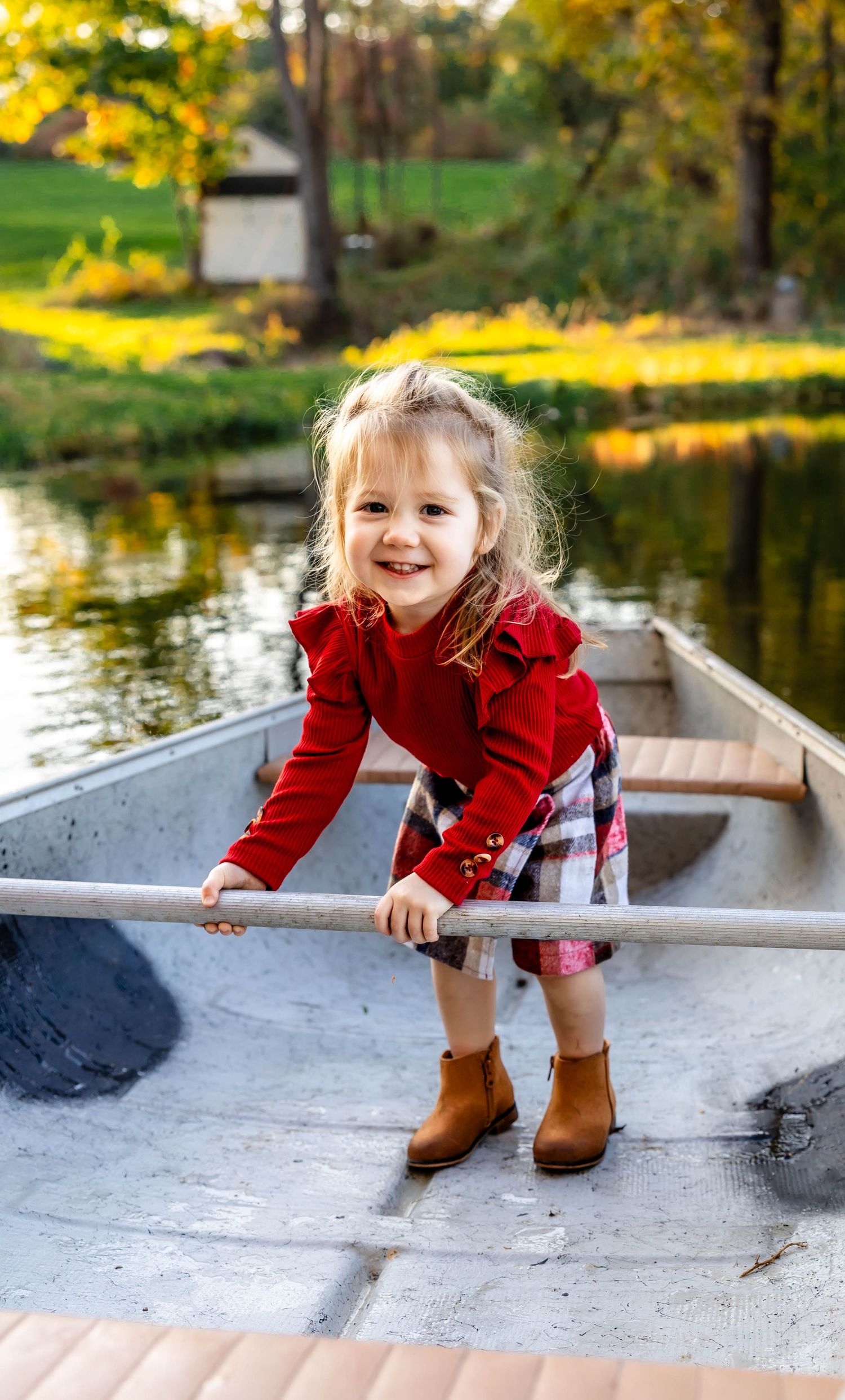 A toddler in a red sweater and brown boots stands playfully in a canoe during autumn.
