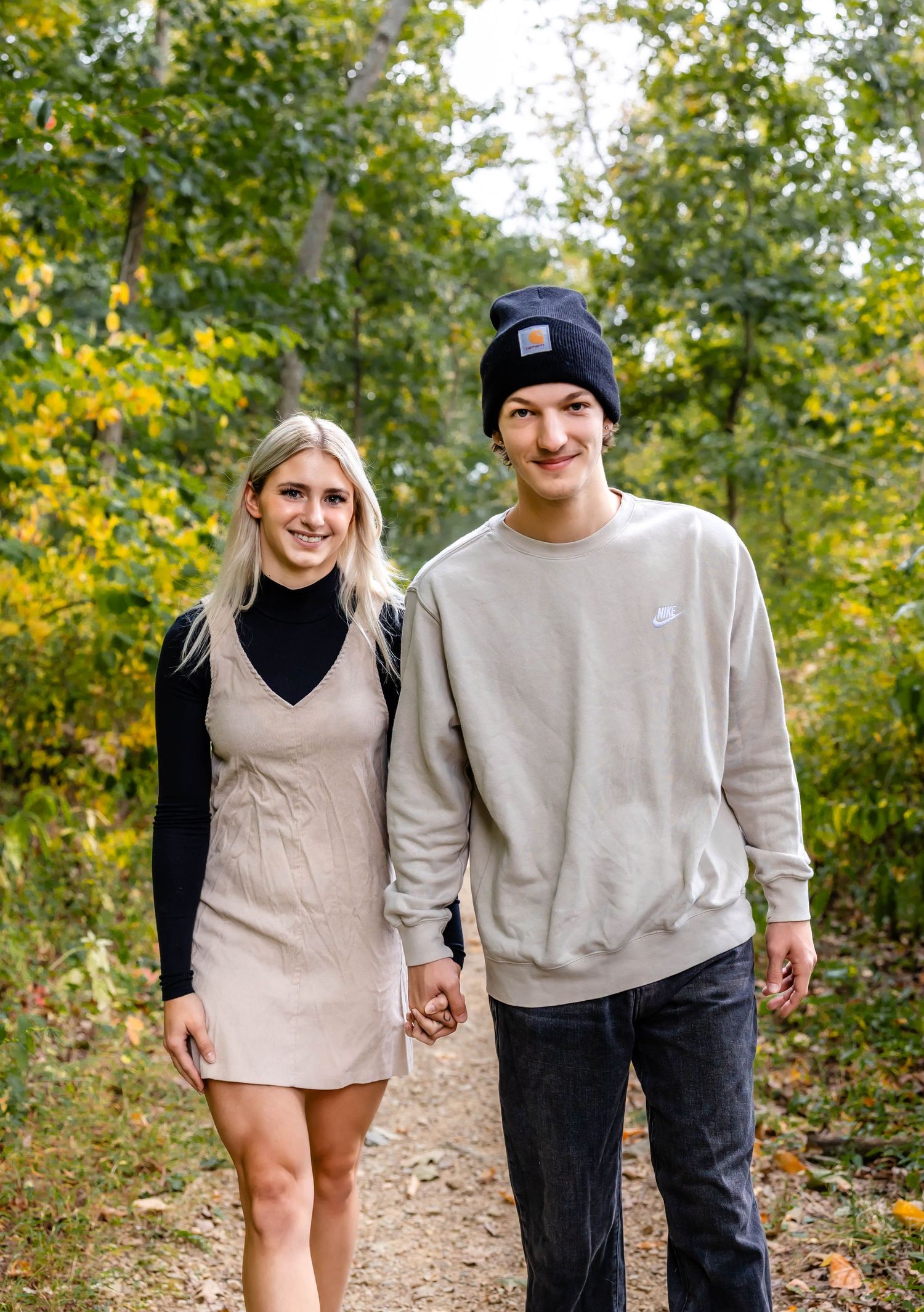 A couple in casual fall attire holding hands while walking on a nature trail surrounded by autumn foliage.