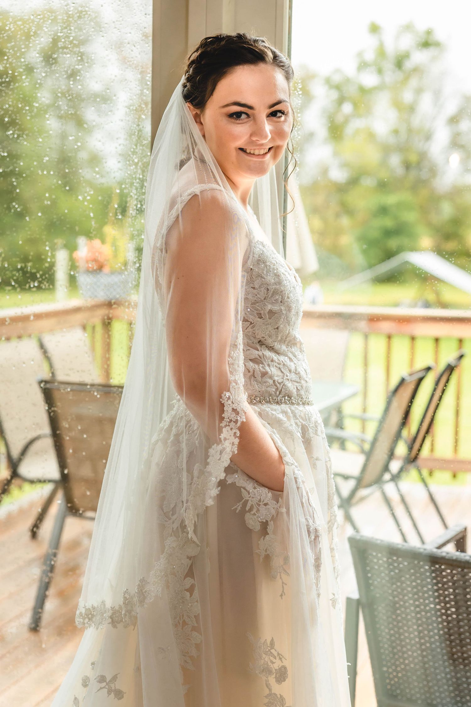 A bride stands by a window in her detailed lace wedding gown and veil.