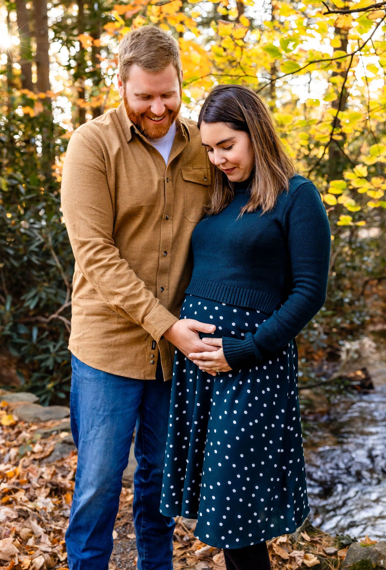 Expectant couple shares intimate moment among fall leaves by stream.