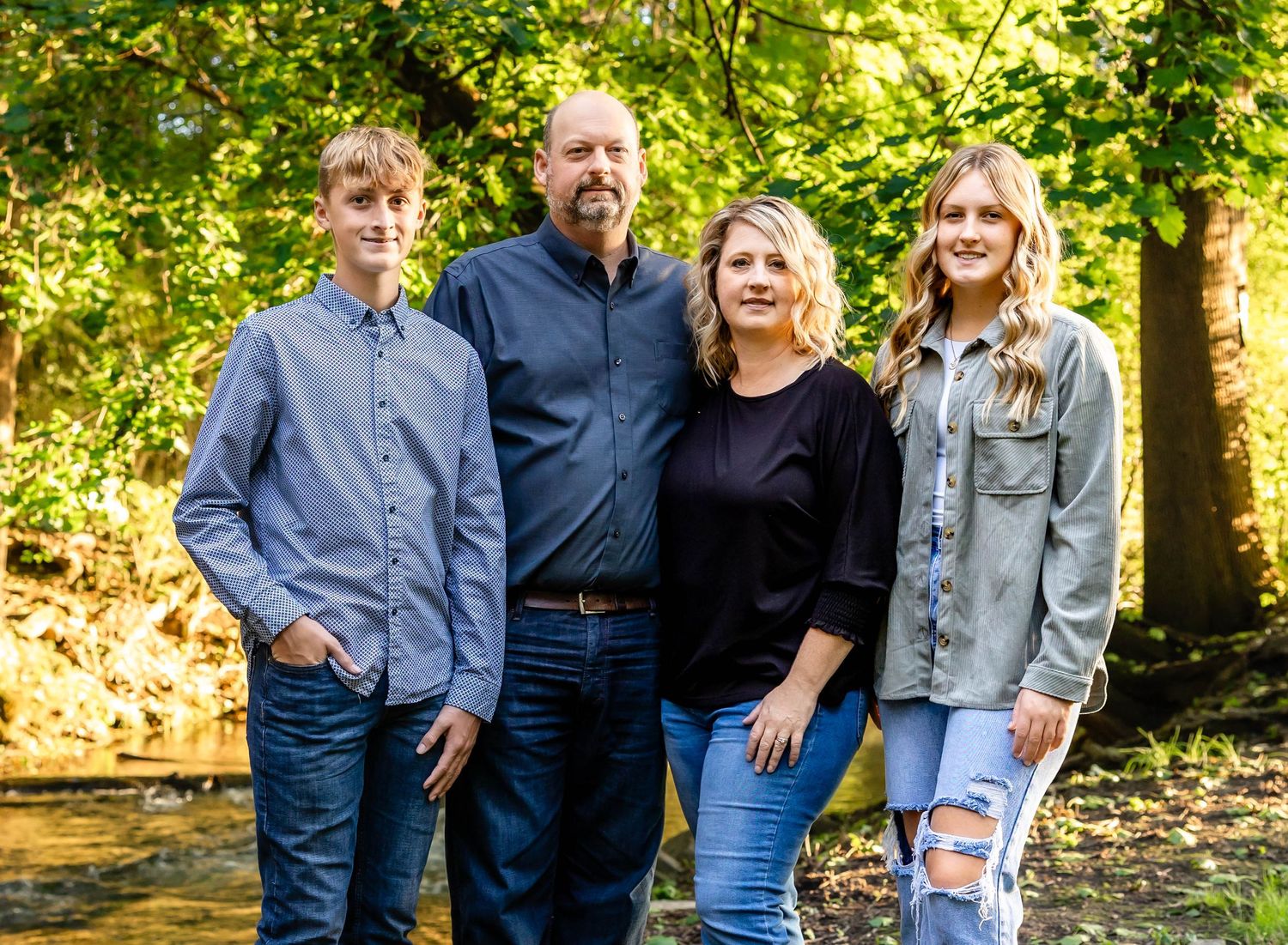 A casual family portrait taken outdoors with greenery and sunlight filtering through the trees.