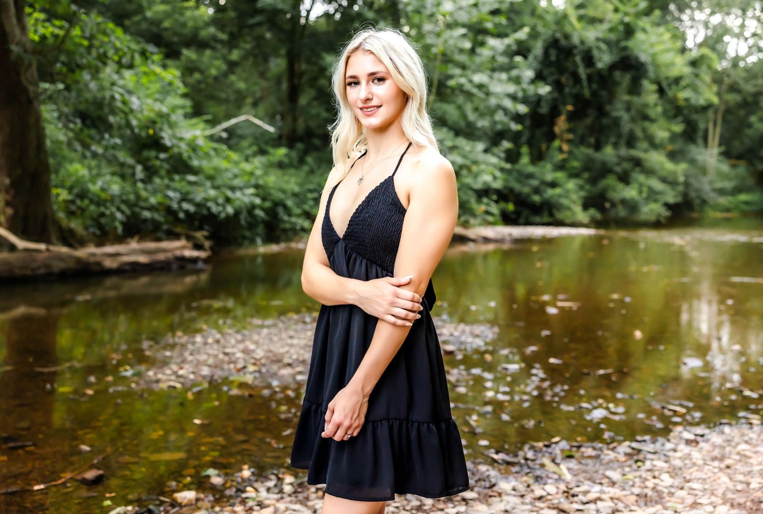 A person in a black sundress poses by a creek in a wooded setting.