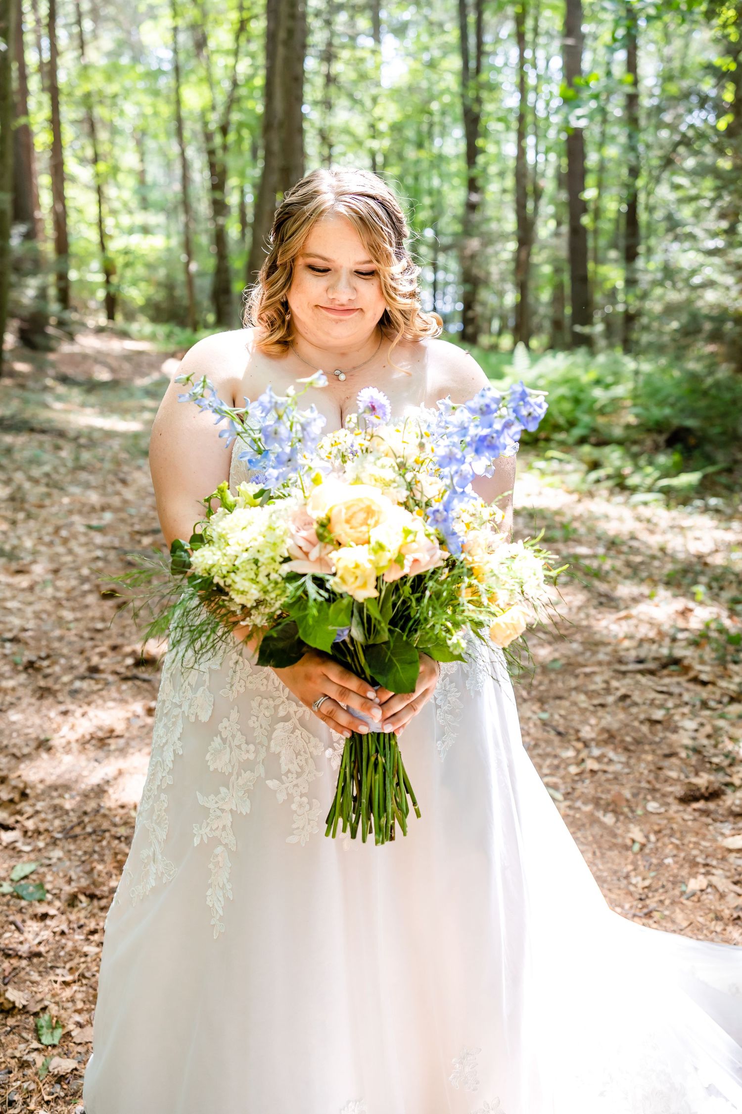 A beaming bride holds a vibrant bouquet of yellow and blue flowers against a forest backdrop.