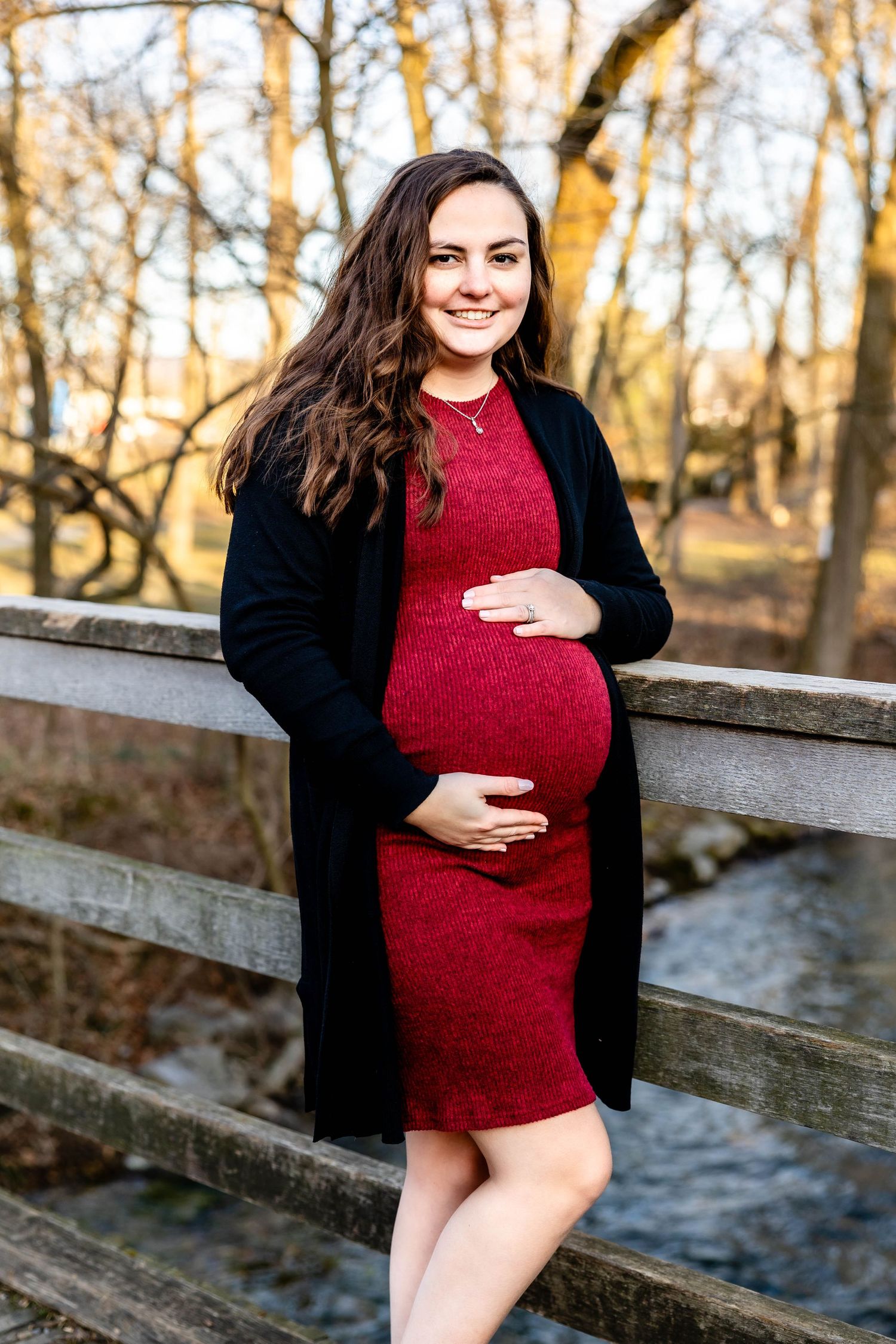 An expectant mother in a red dress and black cardigan poses on a wooden bridge during autumn.