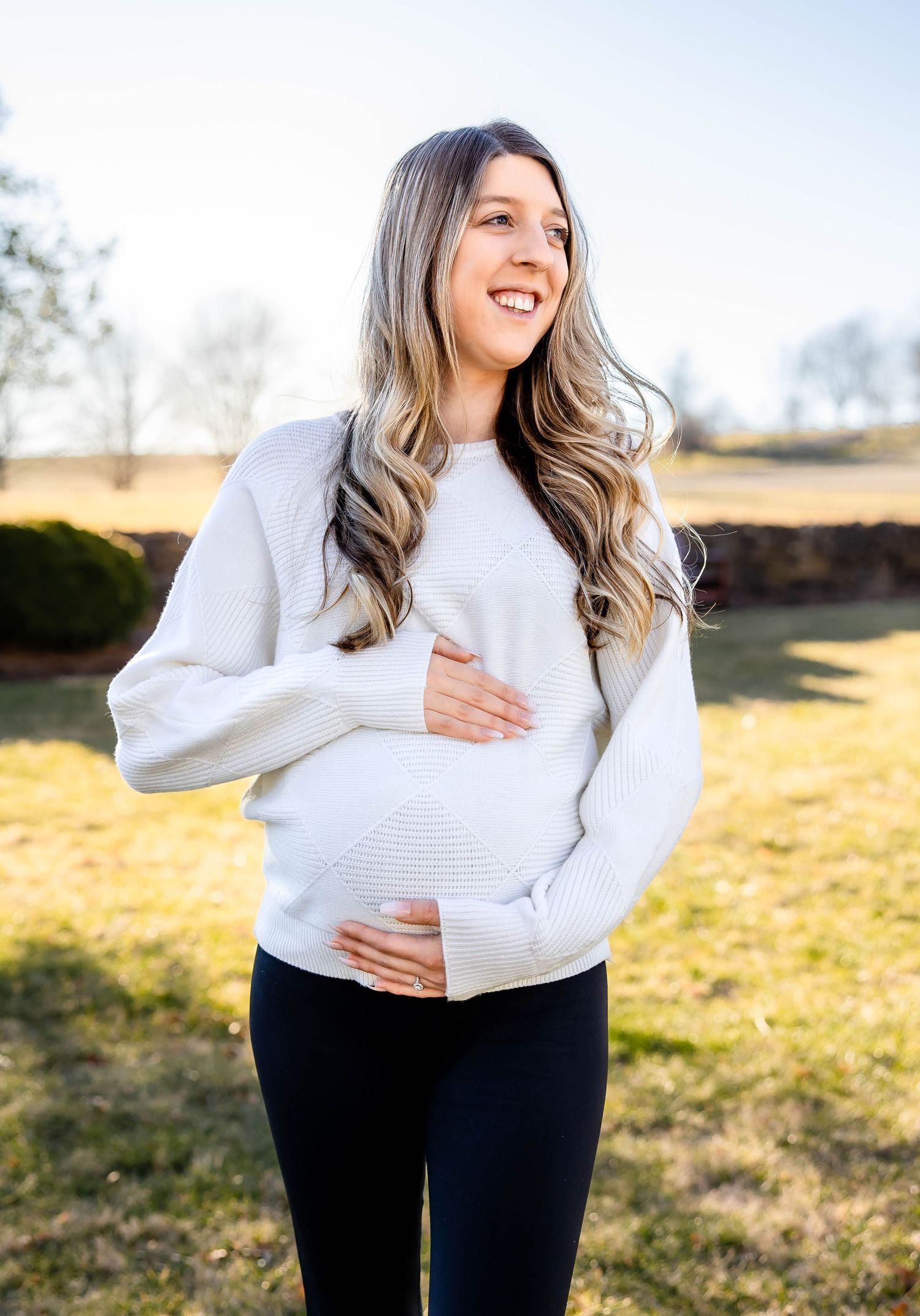 Expectant mother in white sweater and black leggings stands outdoors on sunny day touching her baby bump.