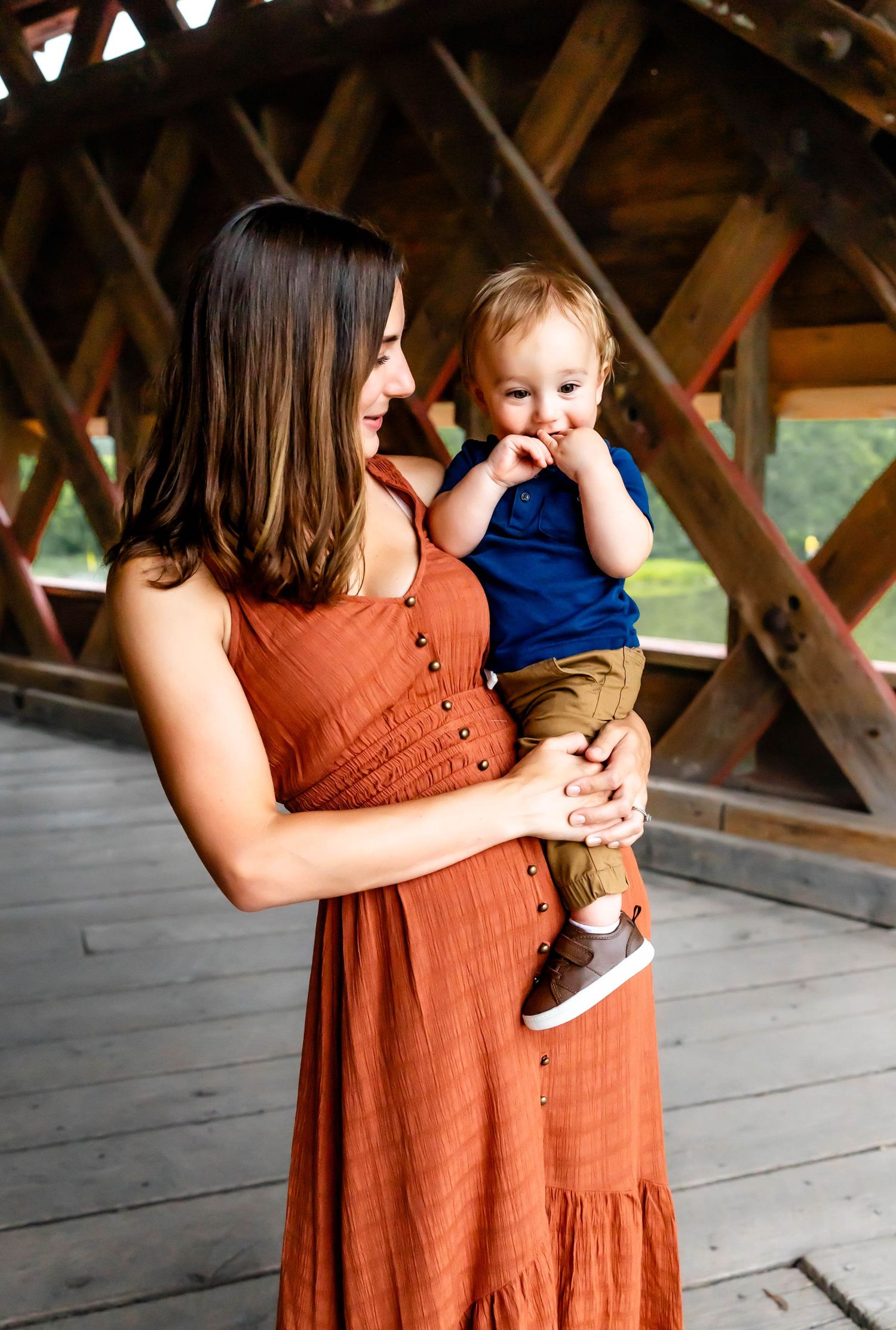 A woman in rust colored dress holds a small child while standing in a wooden covered bridge.