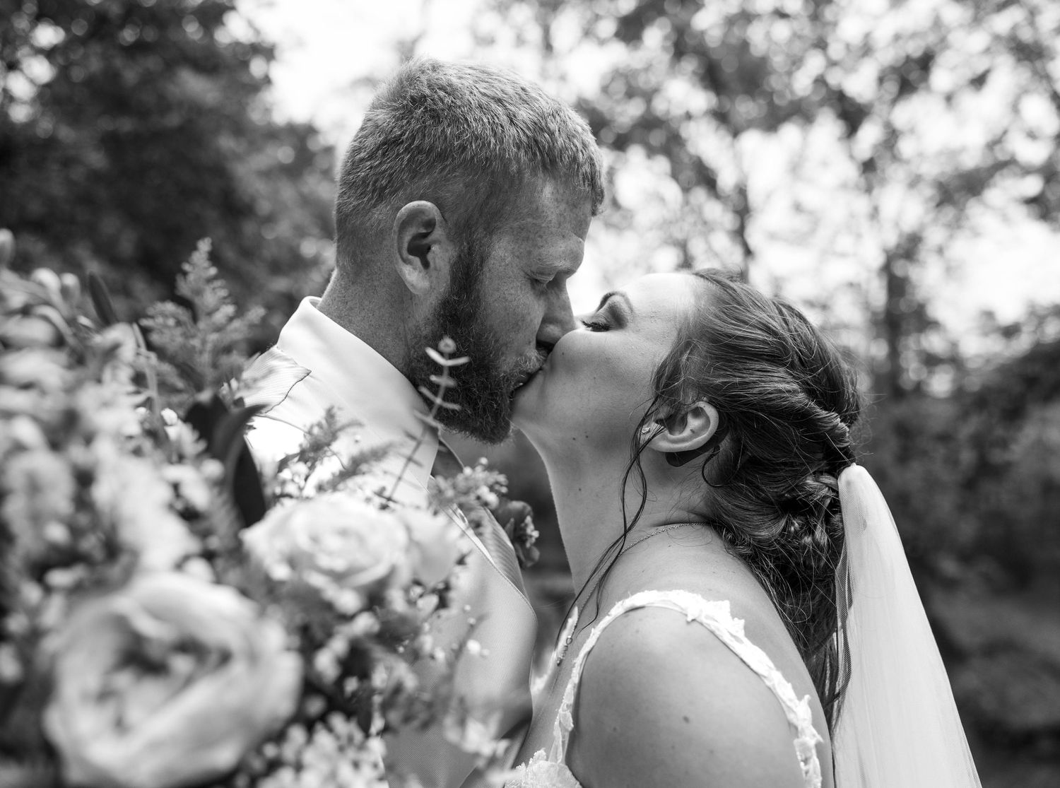 A romantic black and white wedding photo showing a couple kissing outdoors surrounded by flowers.