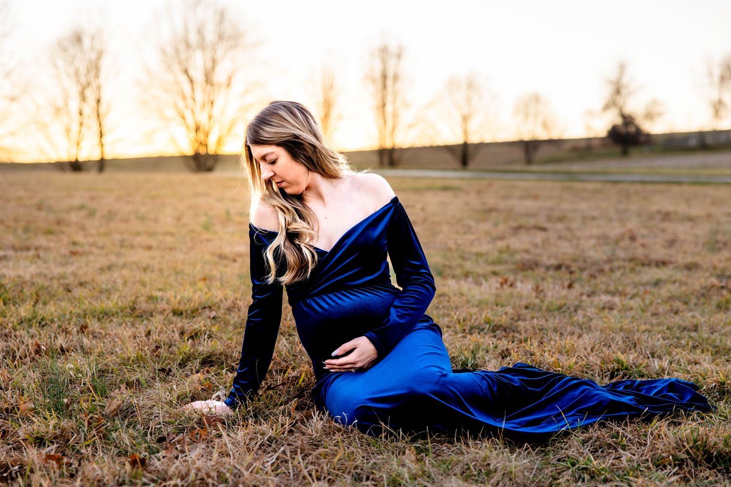 A woman in a blue dress sits gracefully in a field during golden hour.