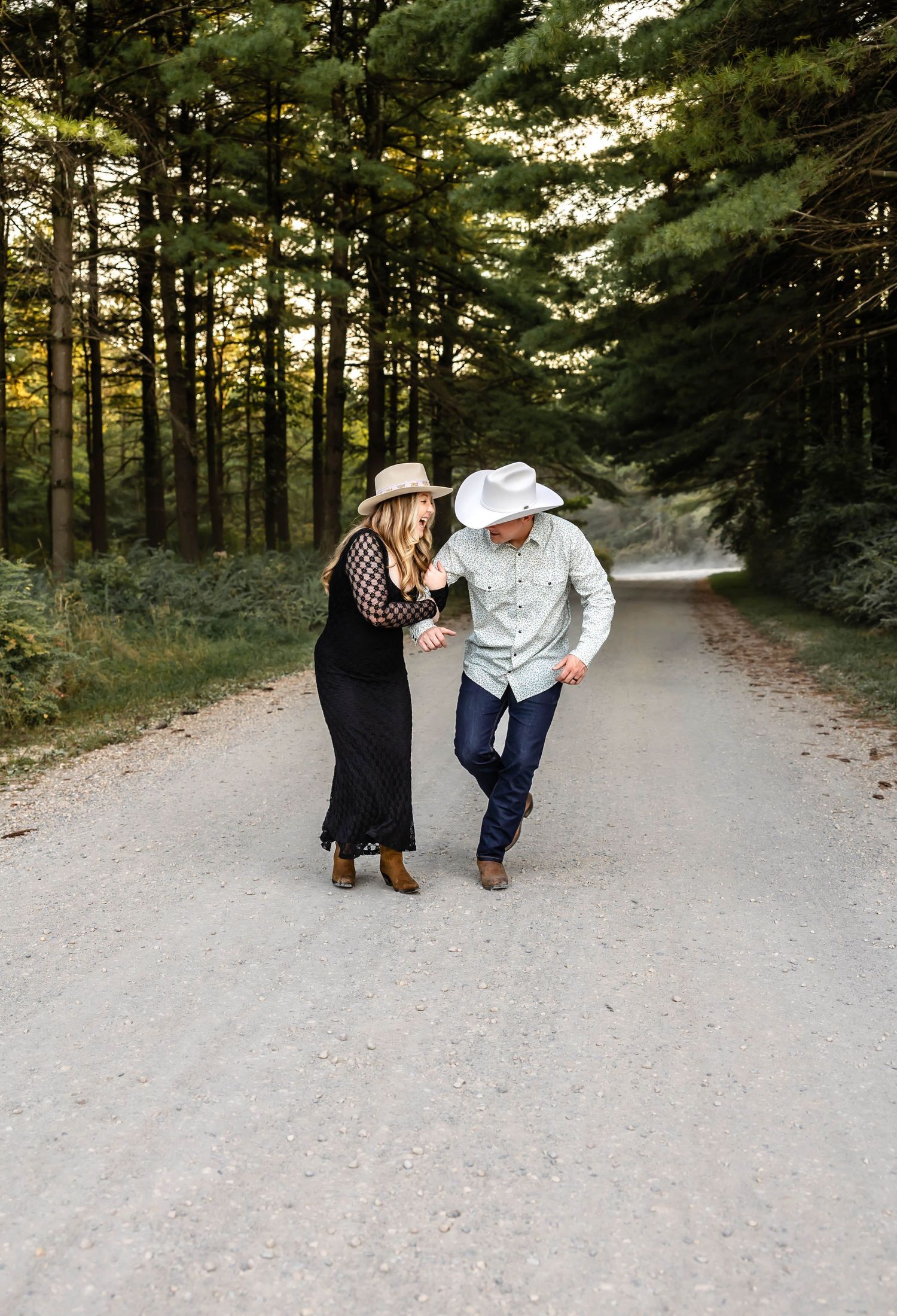 A couple in western attire sharing a lighthearted moment while walking on a forest path.