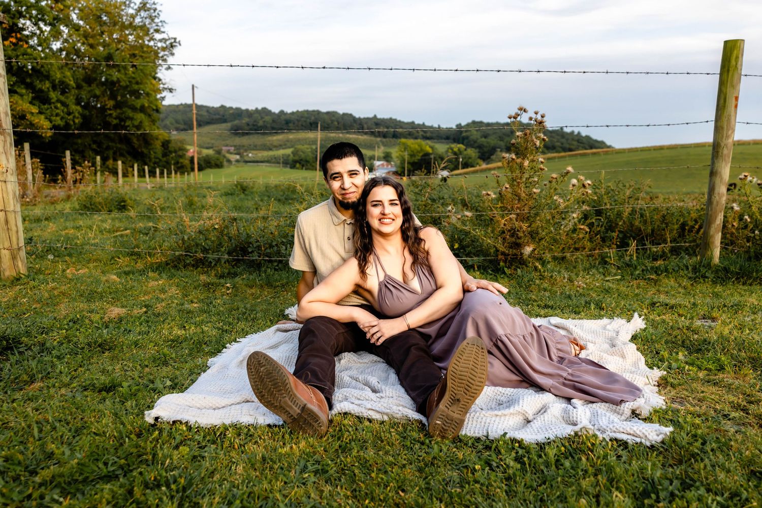 Couple relaxing on a picnic blanket in a scenic countryside setting with rolling hills and fence posts.
