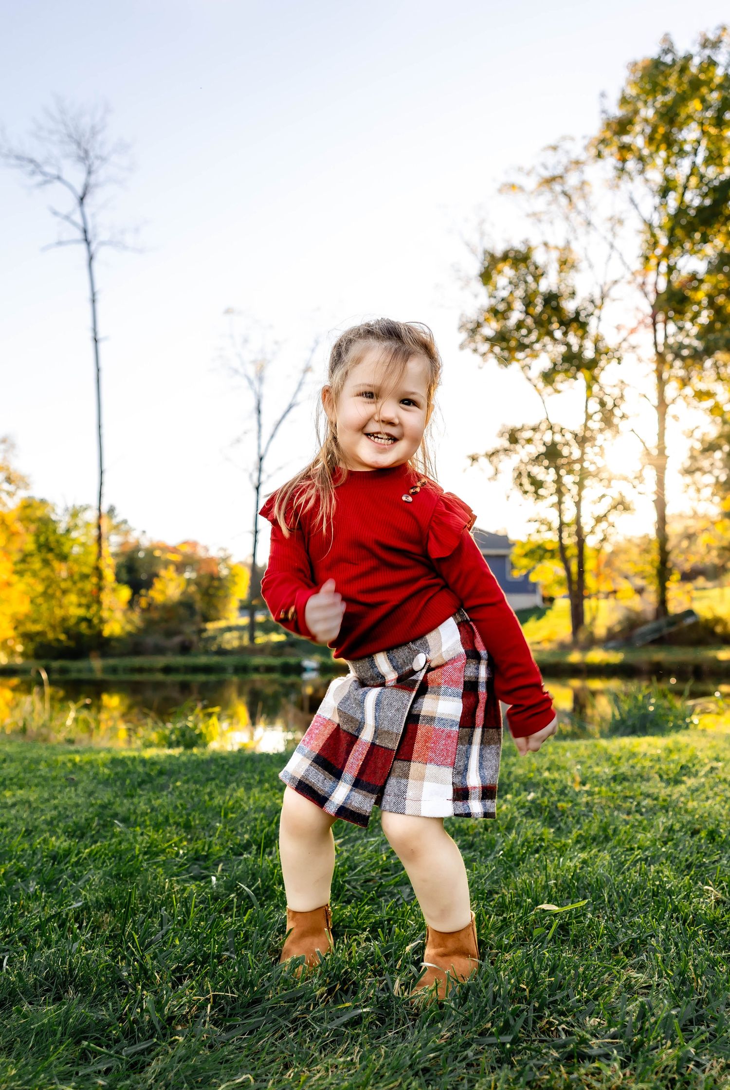 Energetic child wearing red sweater and plaid skirt dances on grass with fall colors in background.