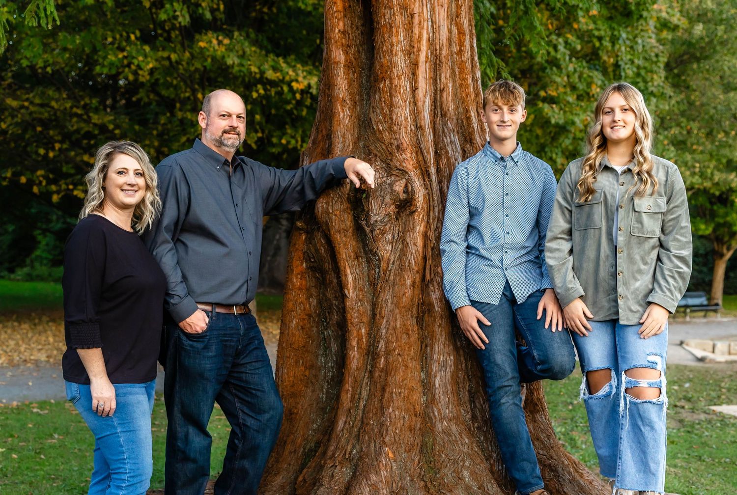 A group poses casually against a large tree trunk in a park setting wearing casual denim and earth-toned clothing.