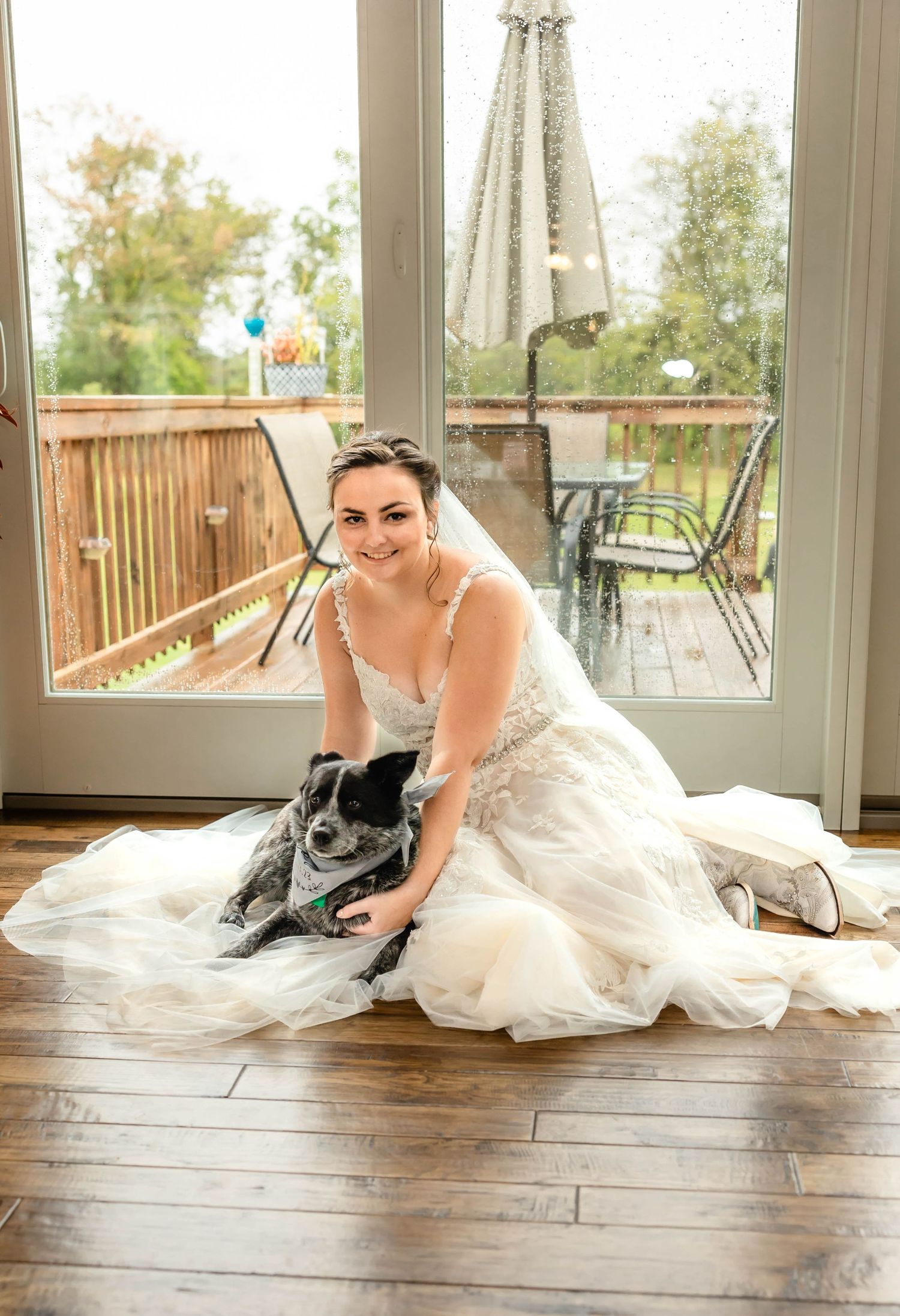 A bride in a white lace dress sits on hardwood floors near glass doors with a gray dog.