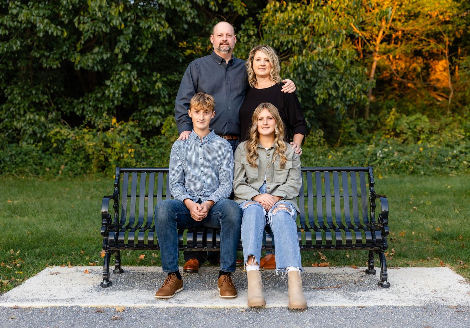 Family of four poses together on green park bench during autumn photo session.