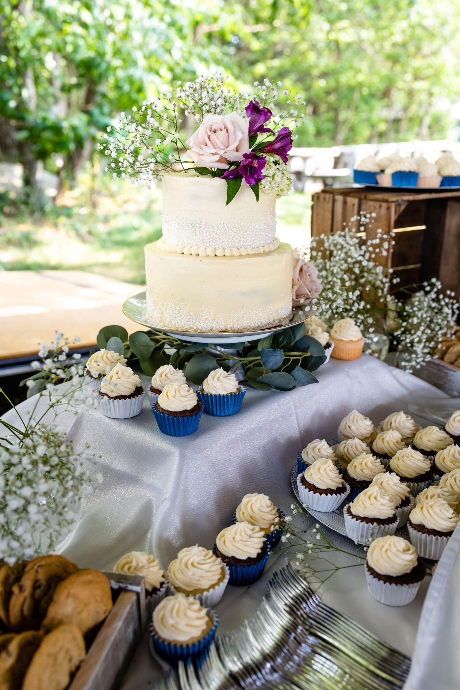 A two-tier white wedding cake with floral decorations surrounded by cupcakes on a dessert table.