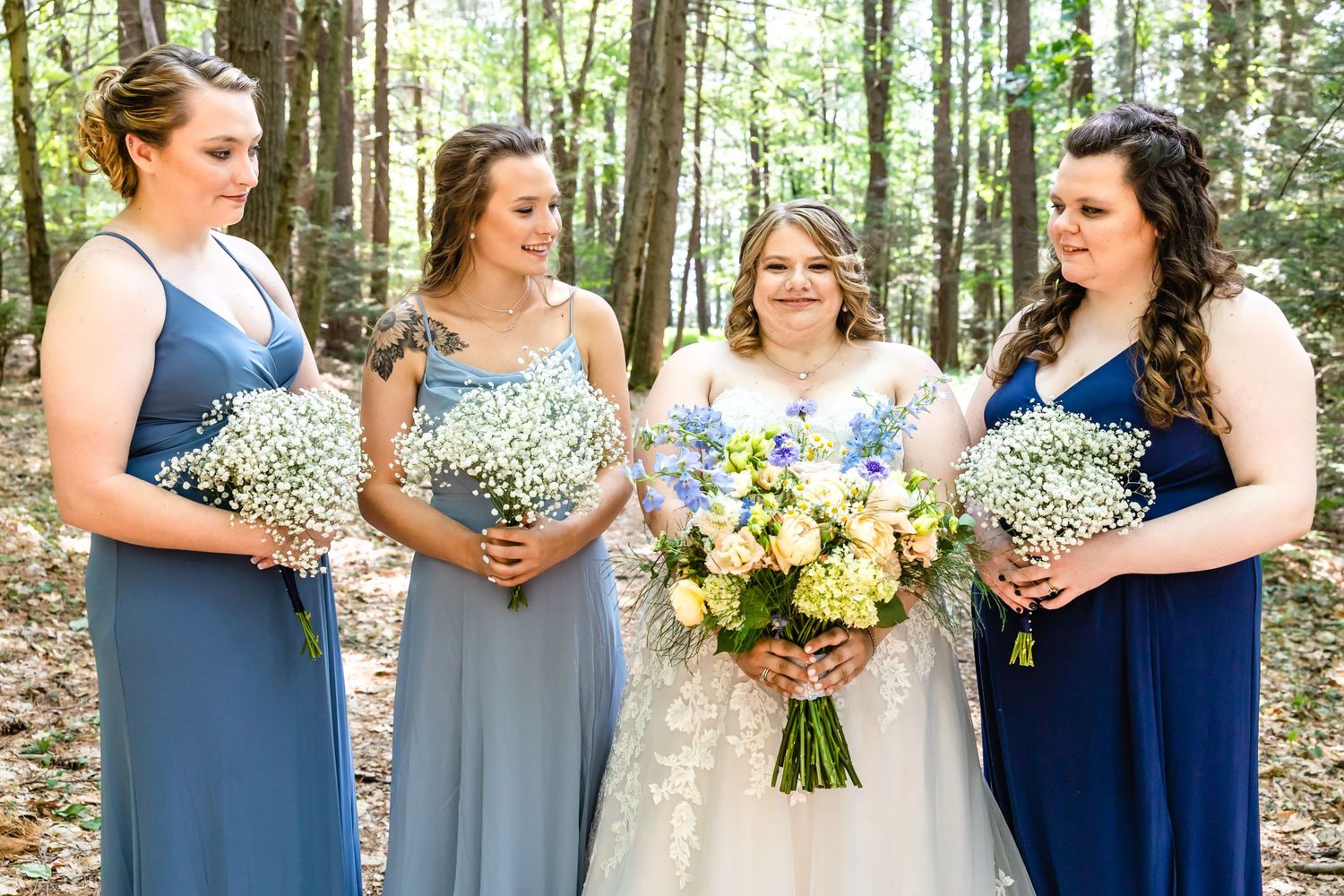 Bridal party in shades of blue holds bouquets while smiling in forest setting.