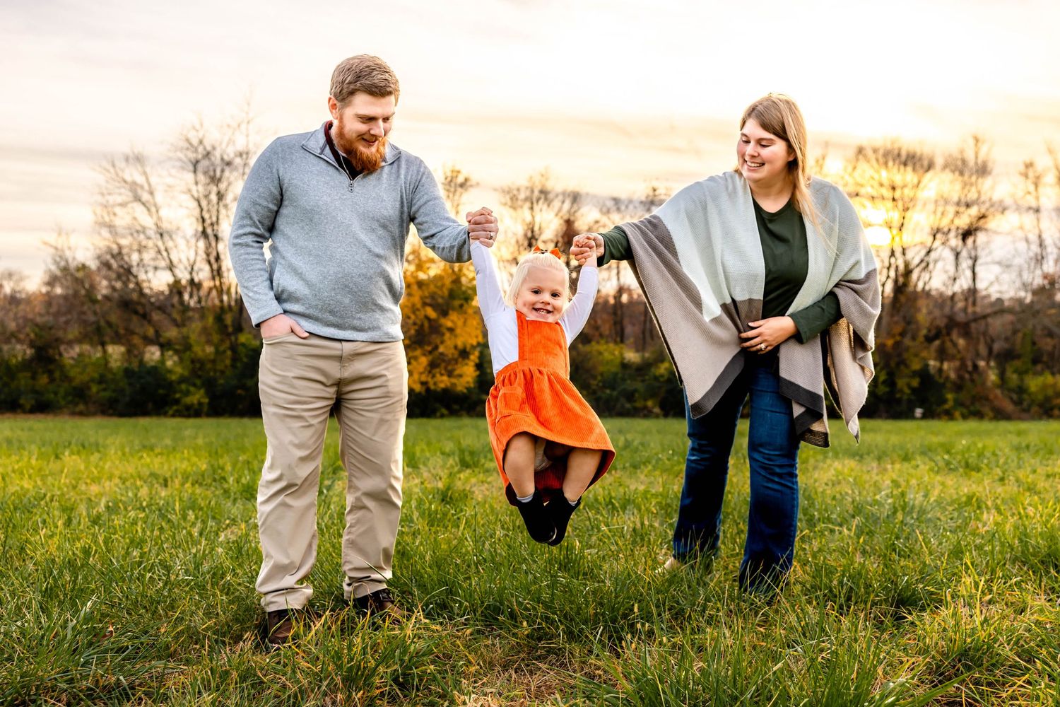 Family plays together in a field during sunset as child jumps holding hands with parents.