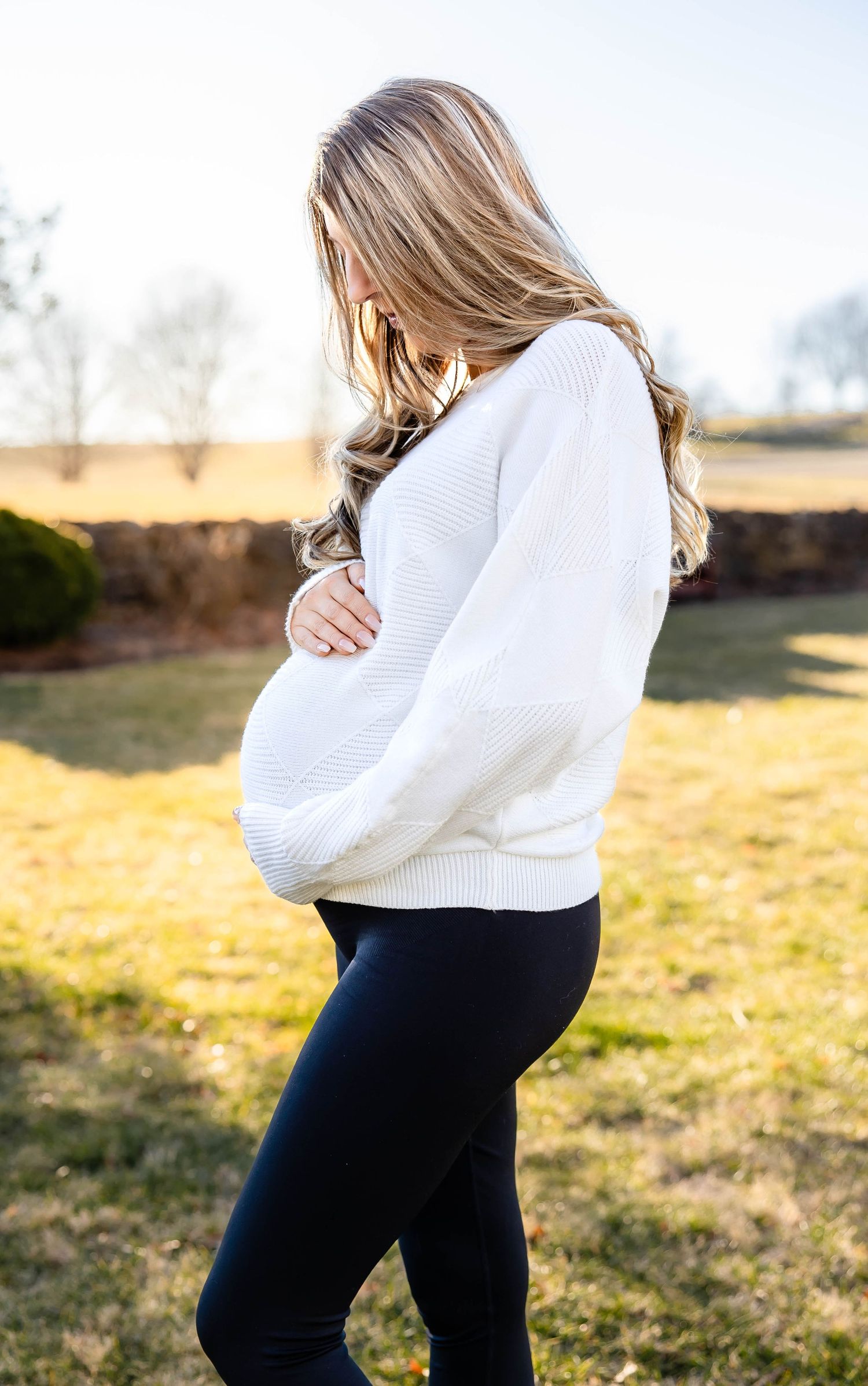 Side profile of pregnant woman in white sweater cradling her belly outdoors in natural setting.