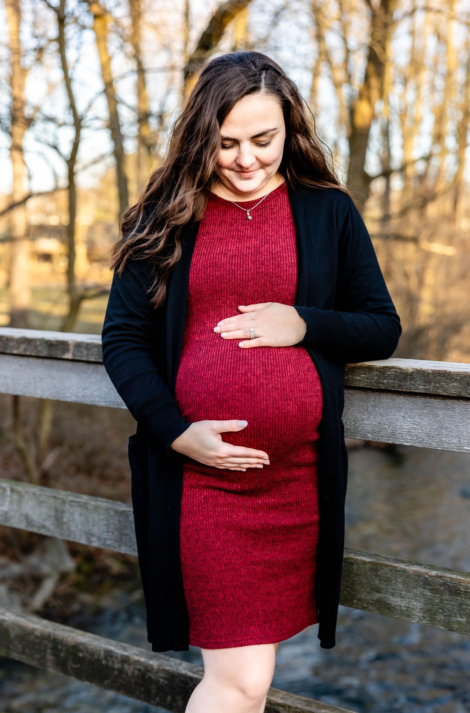 An expectant mother in a red dress touches her belly while standing on a wooden bridge.