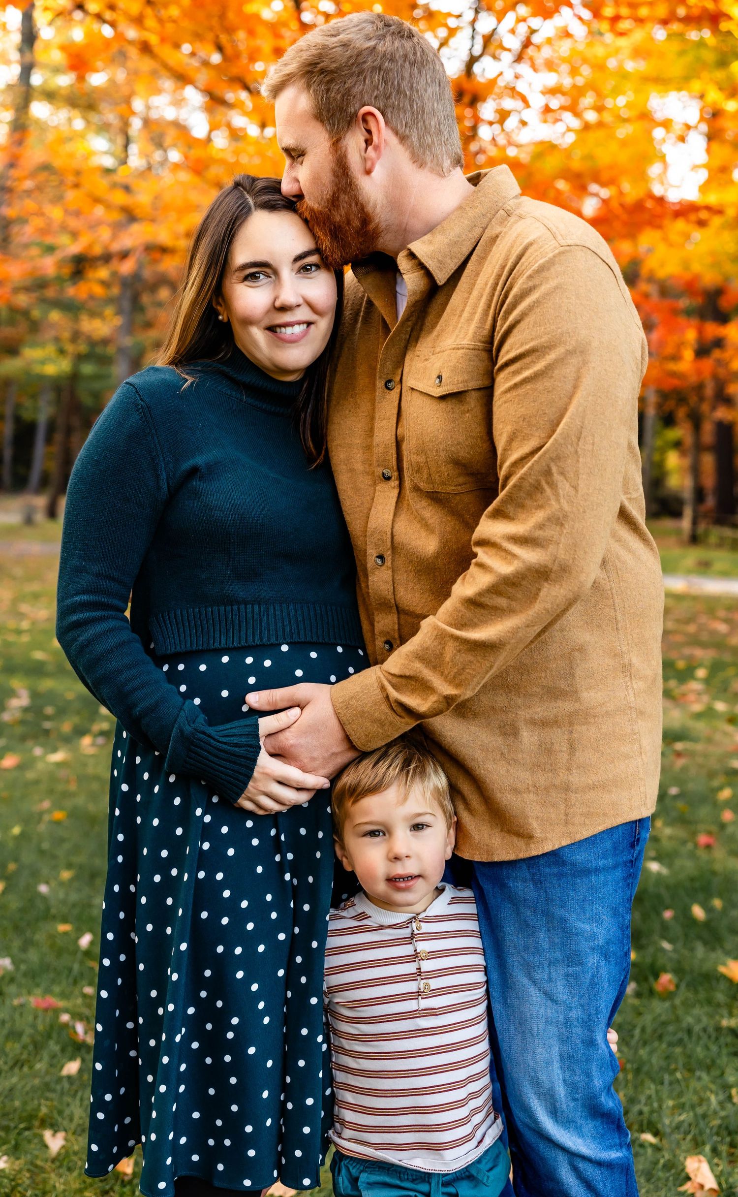 A family shares a tender moment against vibrant fall foliage in an outdoor portrait setting.