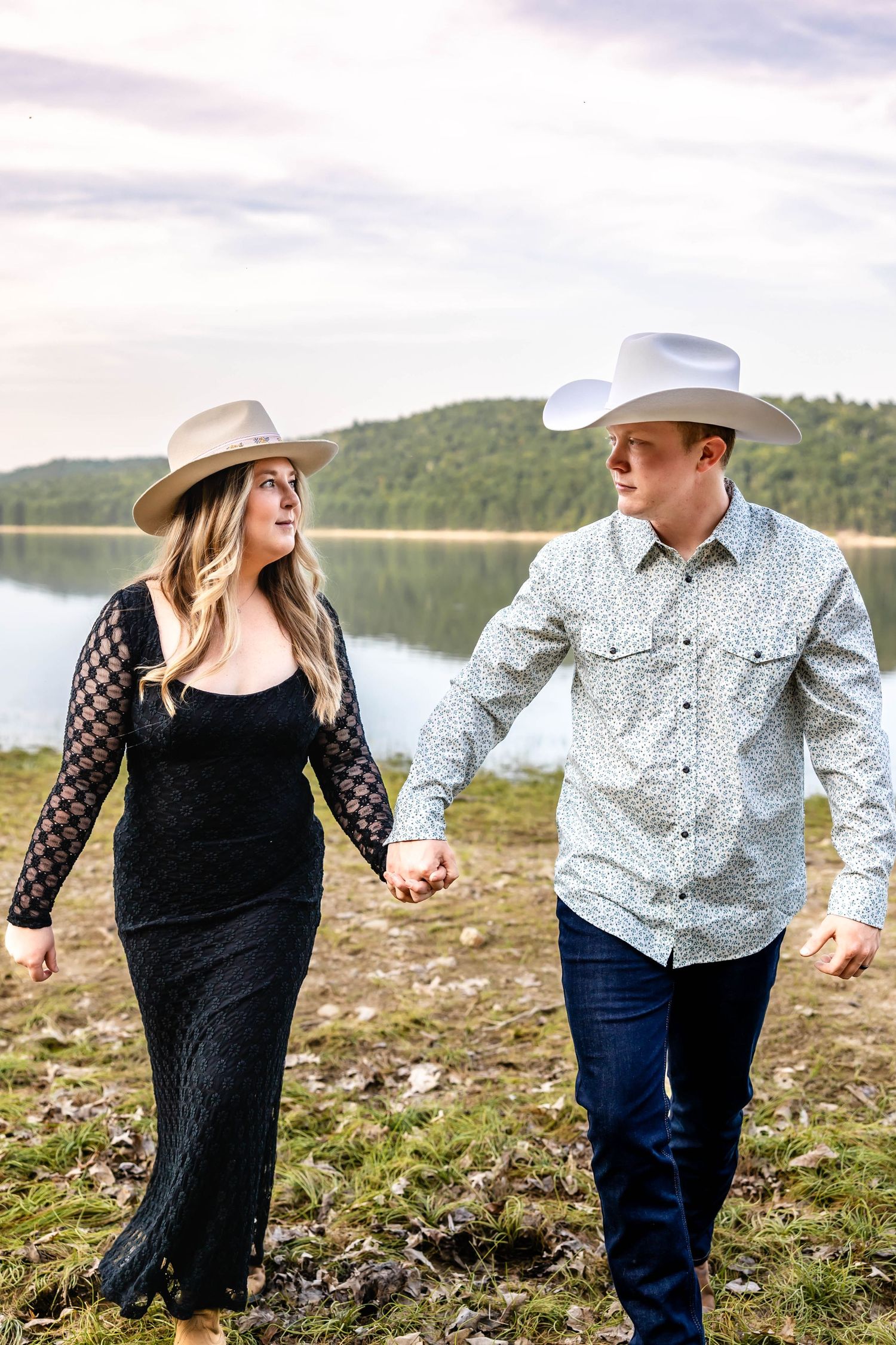 Couple wearing western hats holds hands while walking by mountain lake.