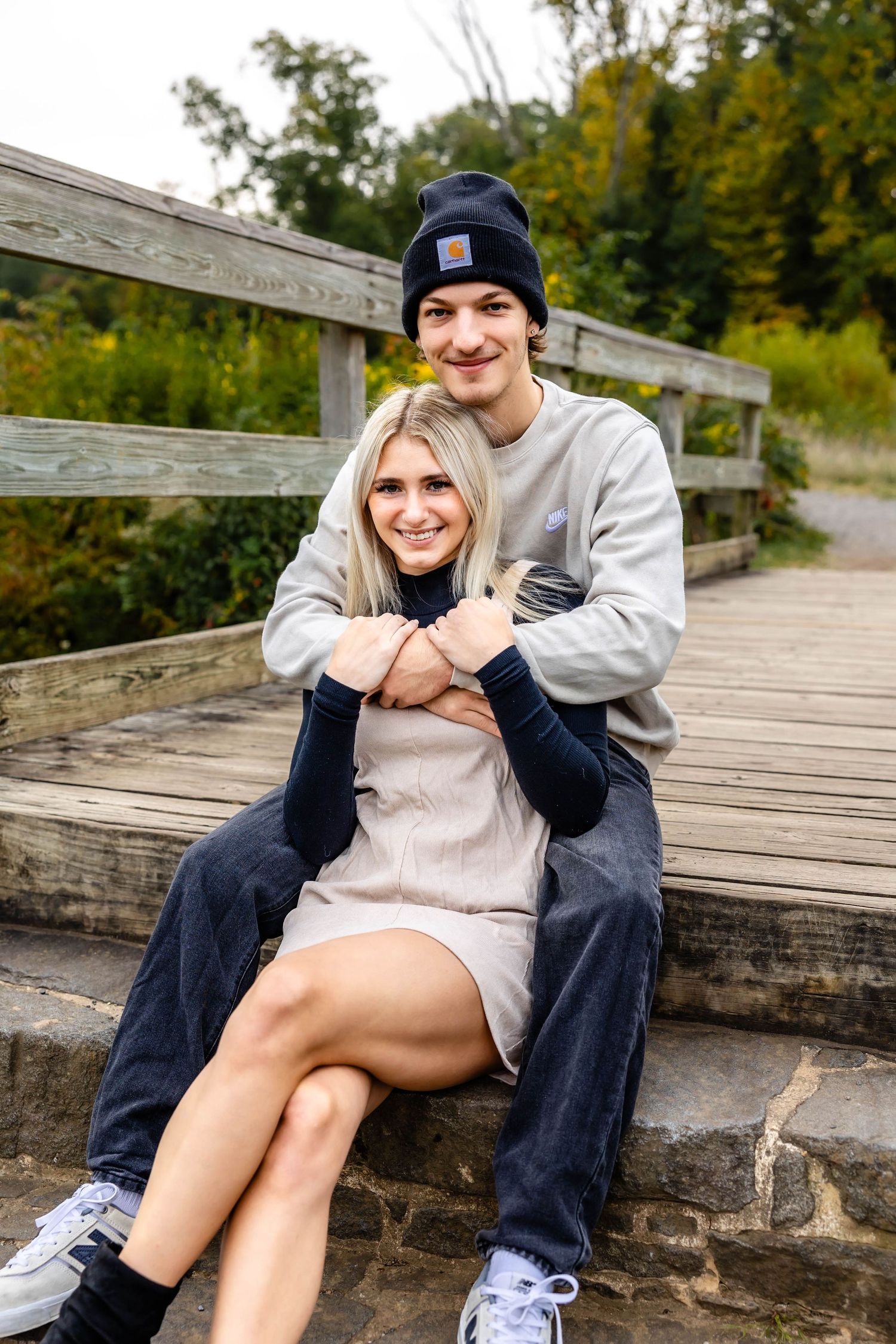 A couple poses together on wooden stairs surrounded by fall scenery.