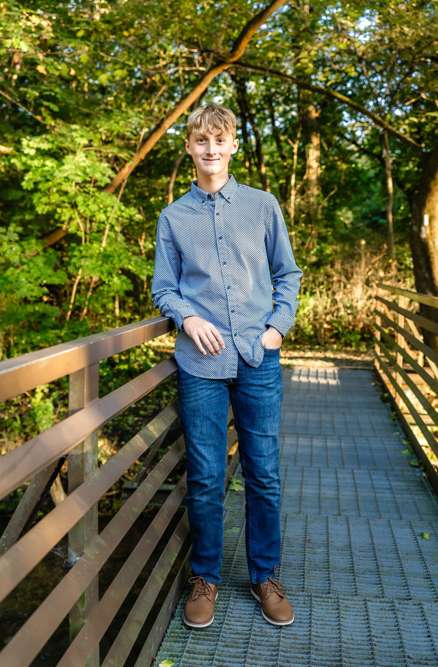 Person in blue button-down shirt and jeans poses on wooden boardwalk in forest setting.