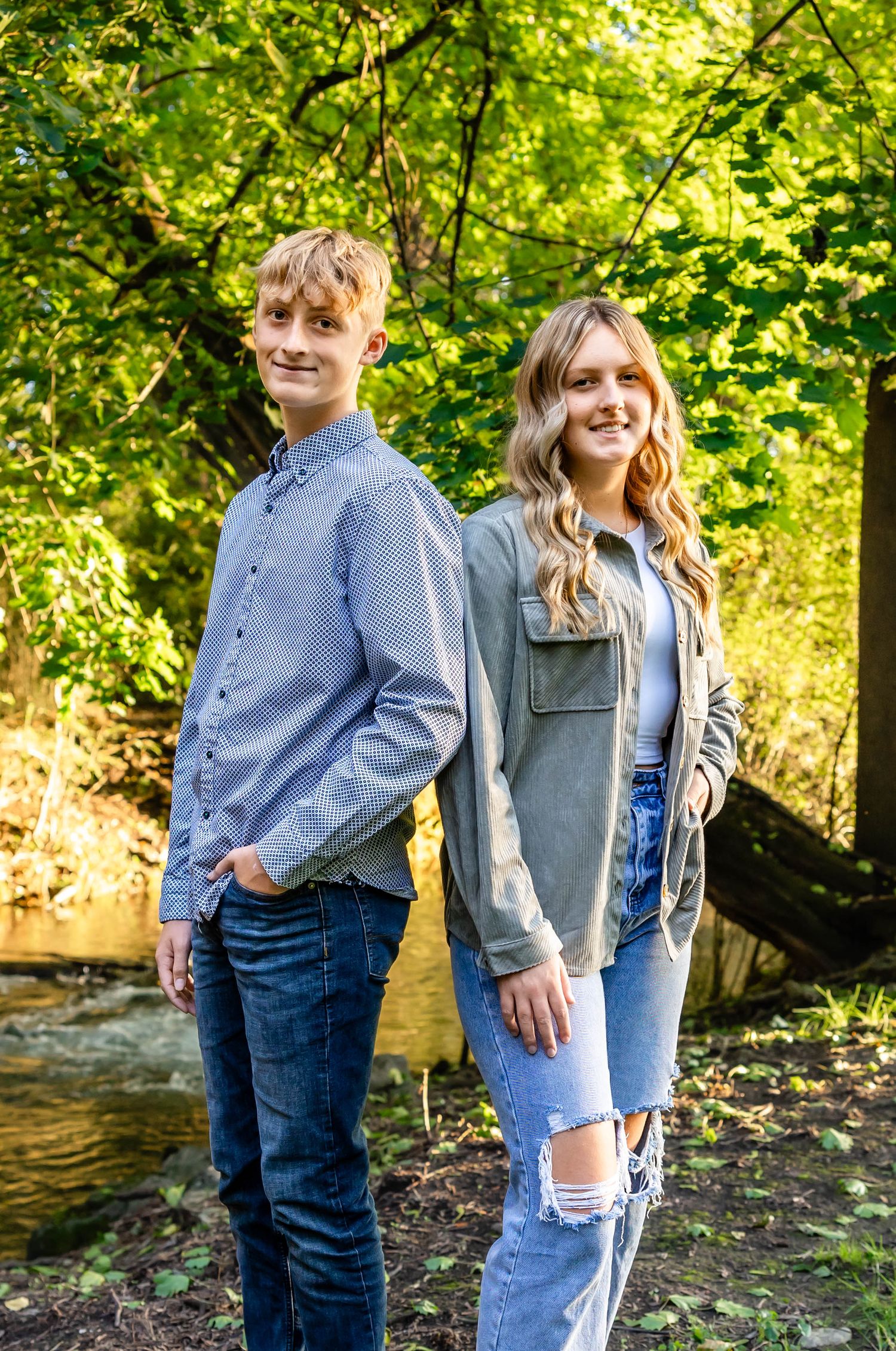 Two teenagers pose casually by a creek with green foliage in the background.