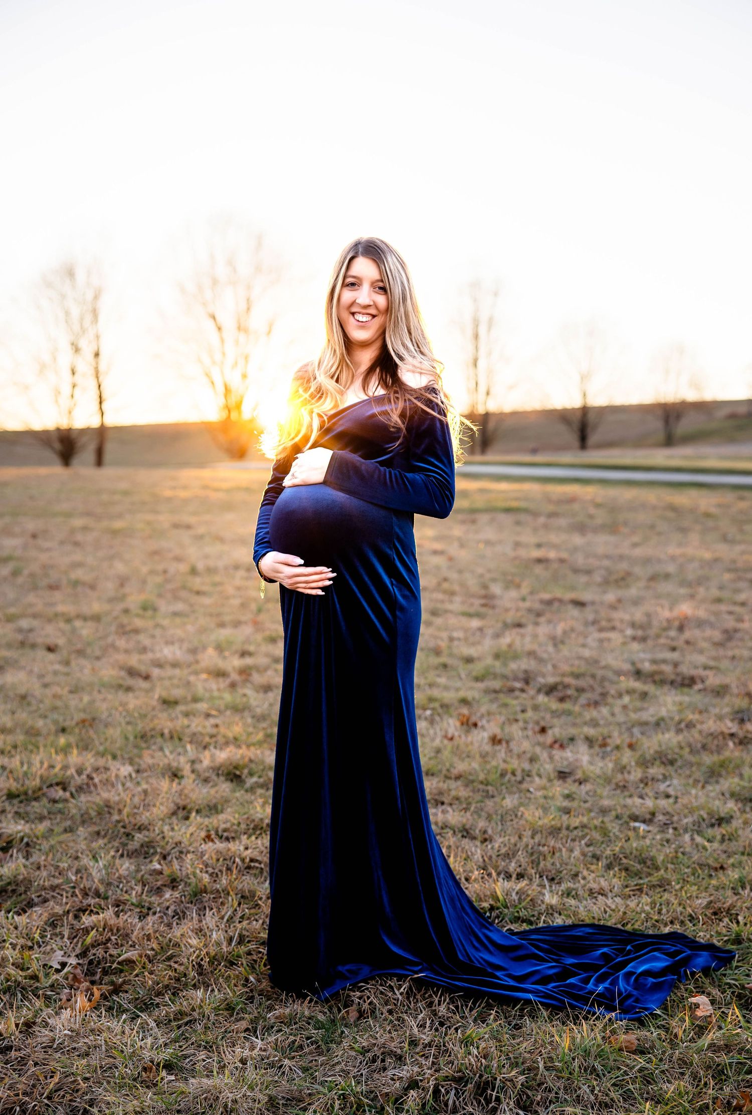 Expectant mother in flowing navy velvet gown poses at sunset in an open field.