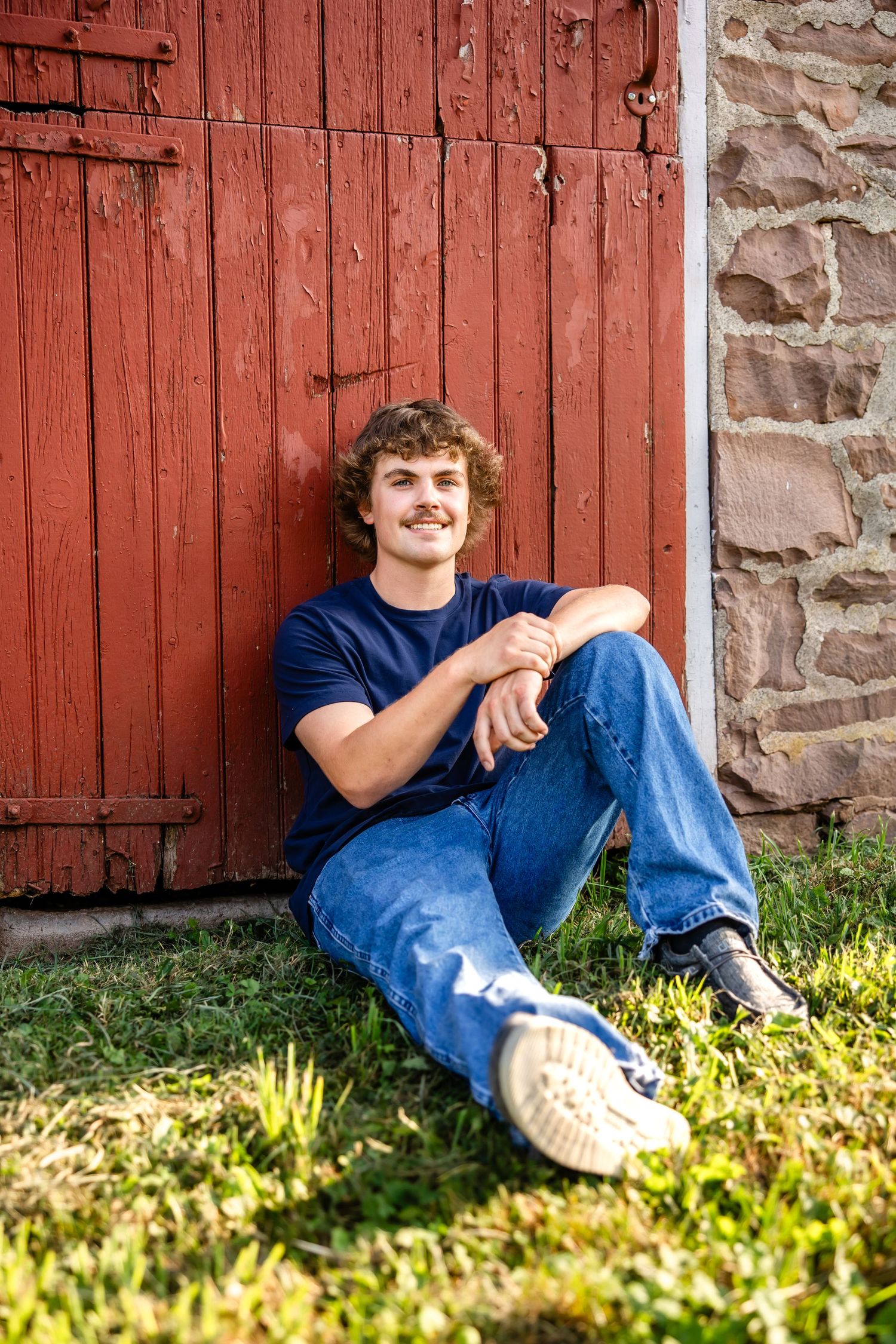 Casual portrait sitting on grass against red barn door in relaxed pose.