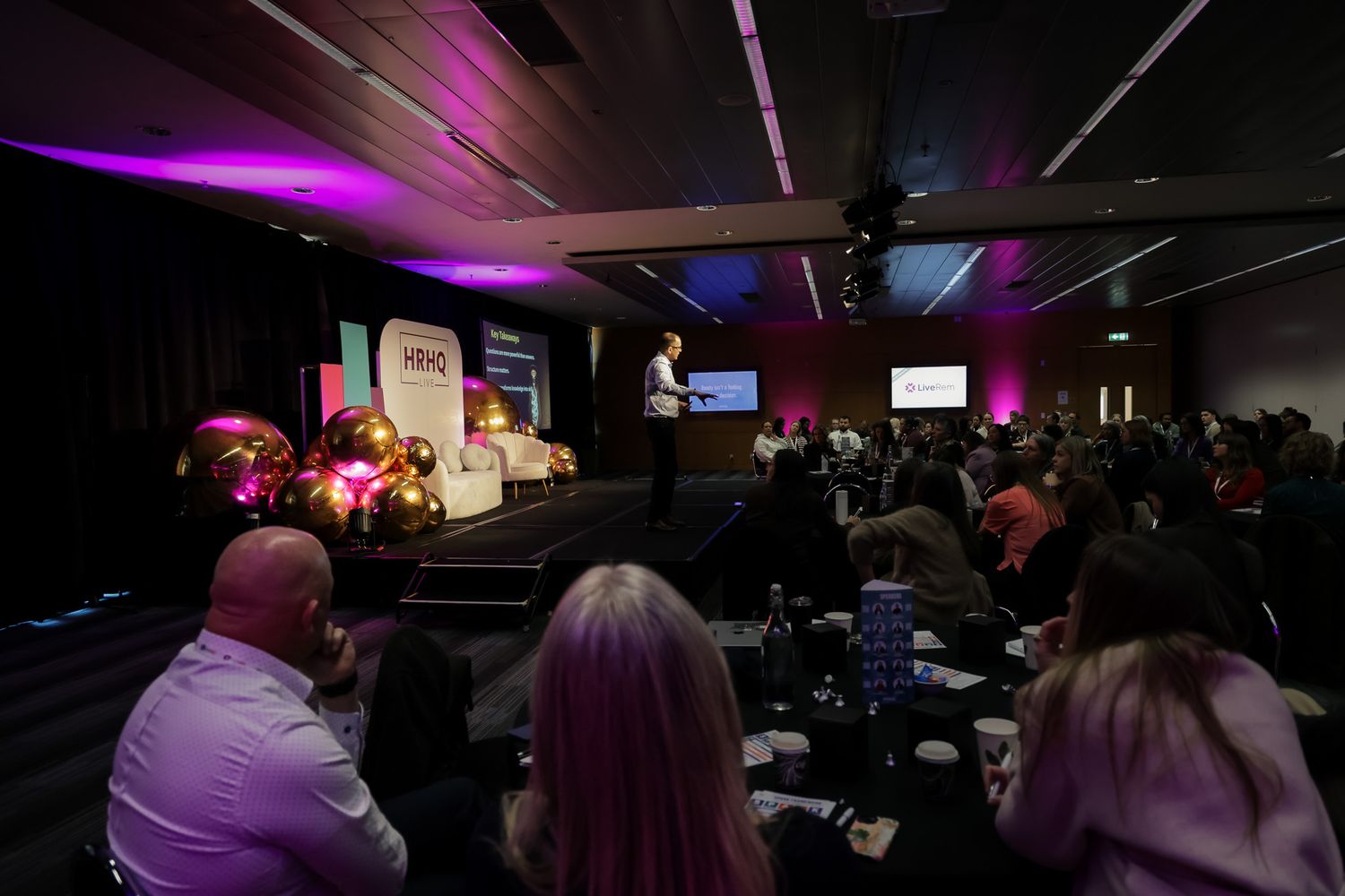 Audience seated at tables during a corporate conference with a speaker on stage under purple lighting.