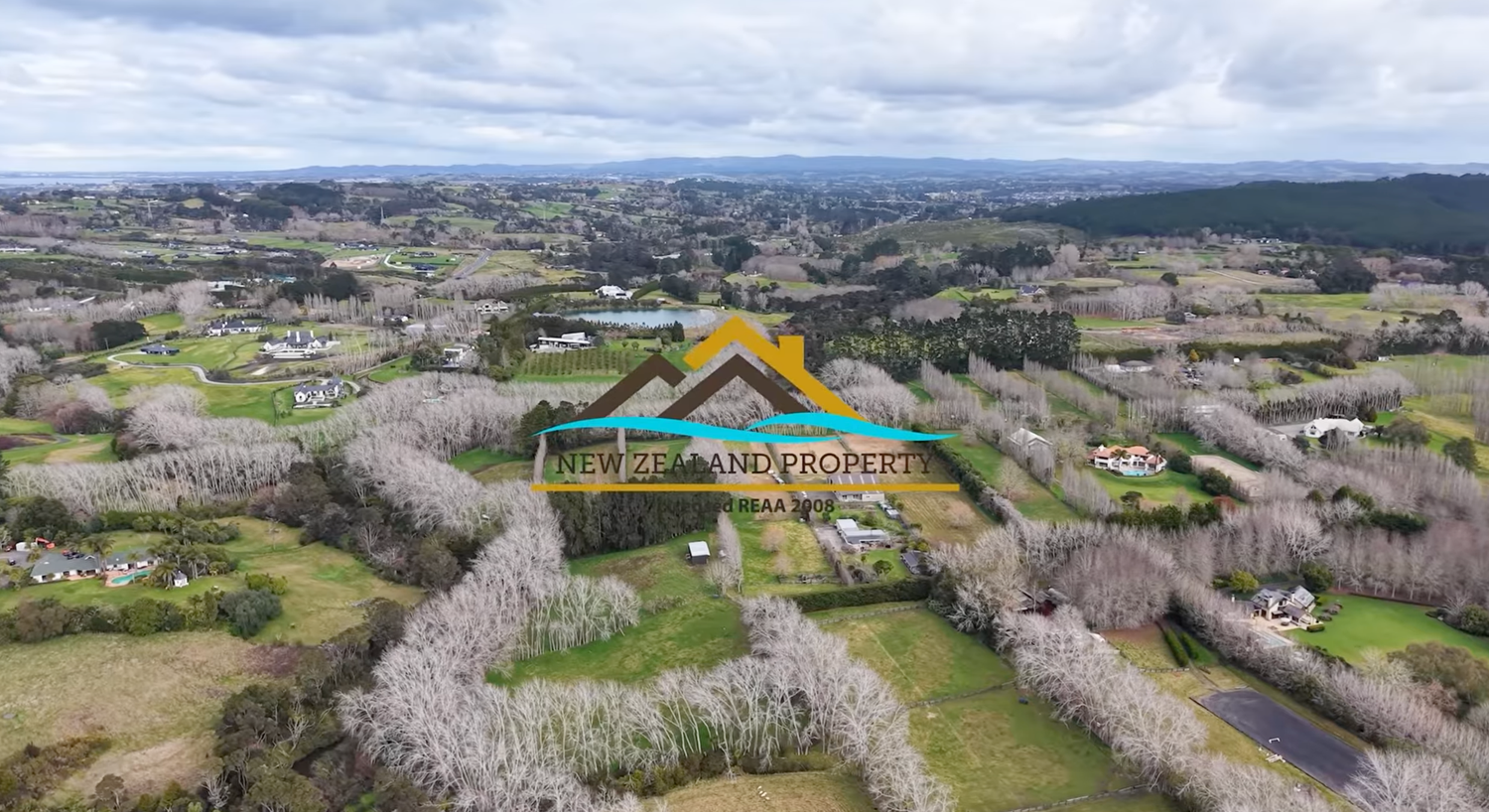 Aerial view of lush green New Zealand countryside with scattered rural properties and tree-lined landscapes under cloudy skies.