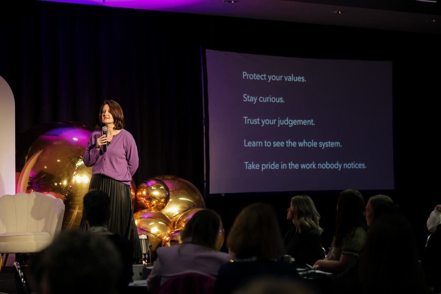 Speaker presenting on stage at conference with motivational values displayed on large screen behind her.