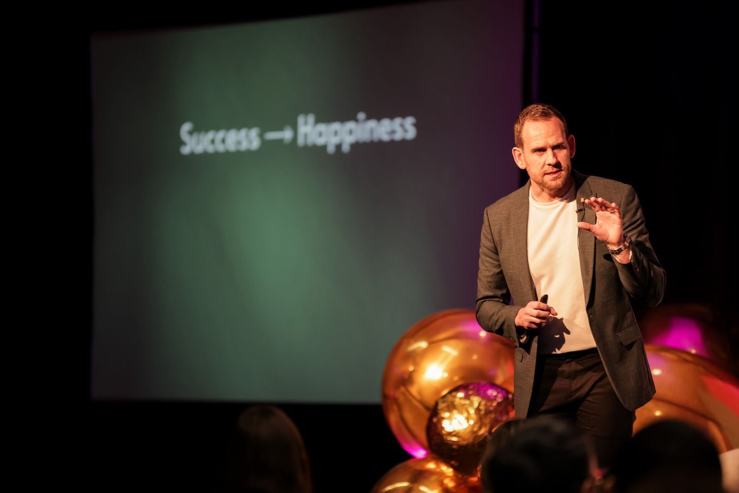 A male speaker presents on stage with a projected slide reading Success equals Happiness behind him at a corporate event.