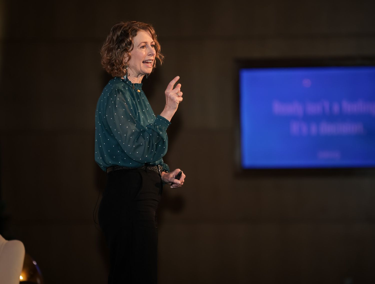 Woman speaker presenting on stage with a blue screen displaying text behind her in a dimly lit venue.