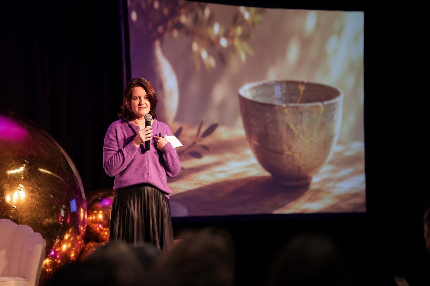 Woman in purple sweater speaks at podium with microphone, large projected image of ceramic bowl on stage behind her.