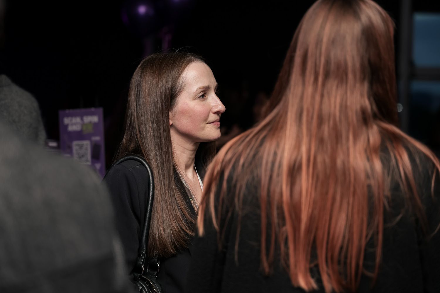 Two women engaged in conversation at a dimly lit social event, one facing forward with straight brown hair.