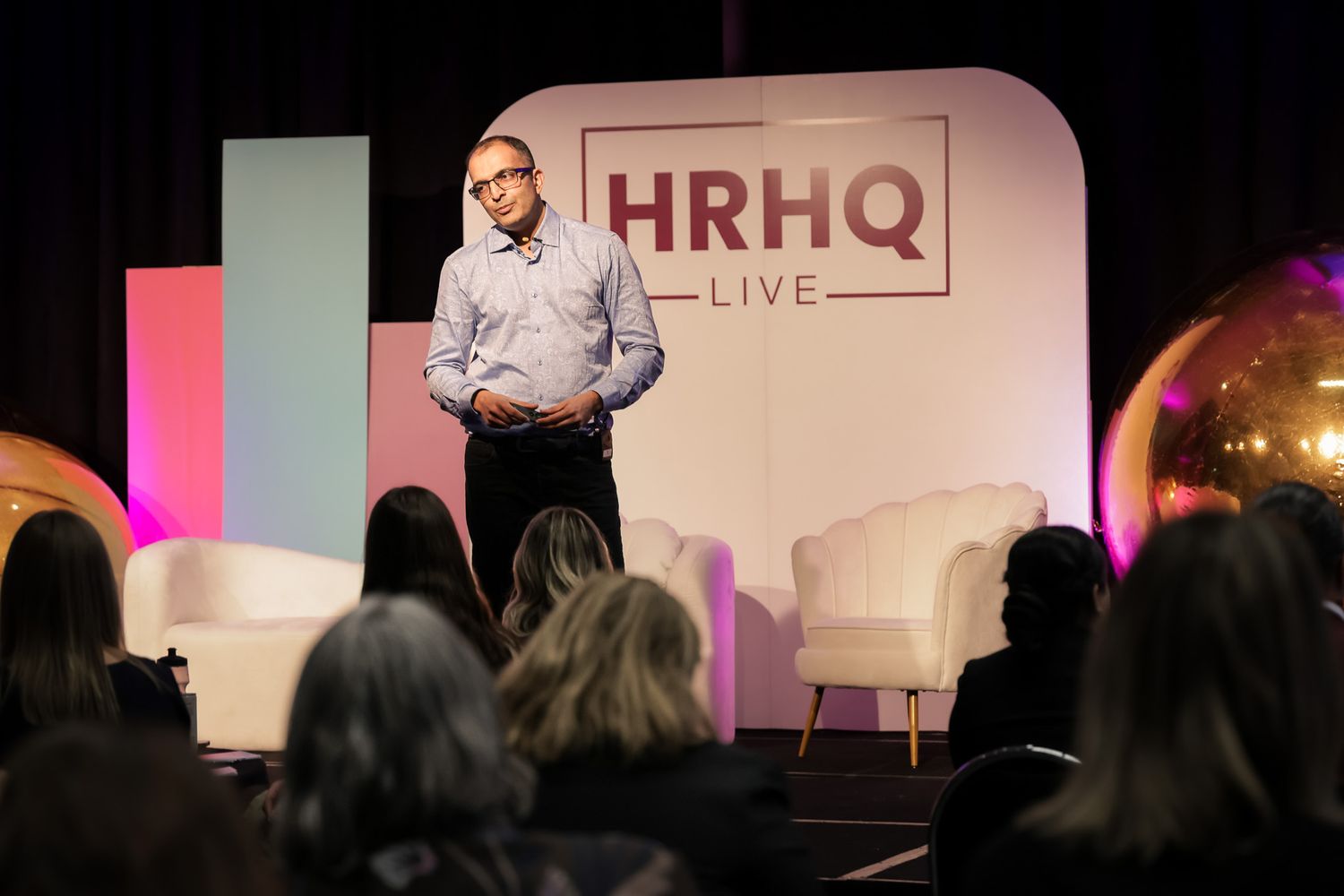 A speaker presents on stage at HRHQ Live event with white chairs and branded backdrop in a dimly lit conference hall.