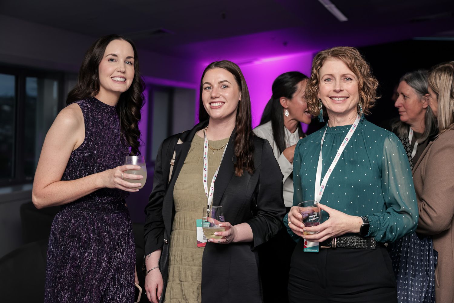 Three women smiling and holding drinks at a professional networking event in a modern venue.