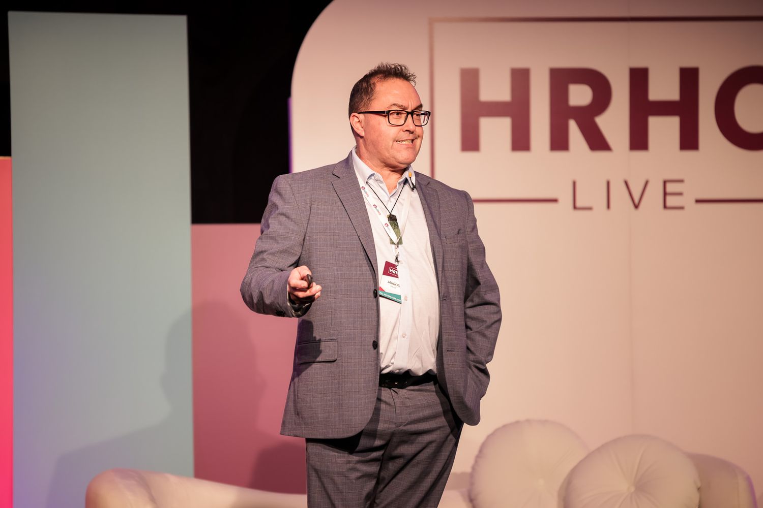 A male speaker presents on stage at HRHOT LIVE conference, wearing a gray blazer under professional lighting.