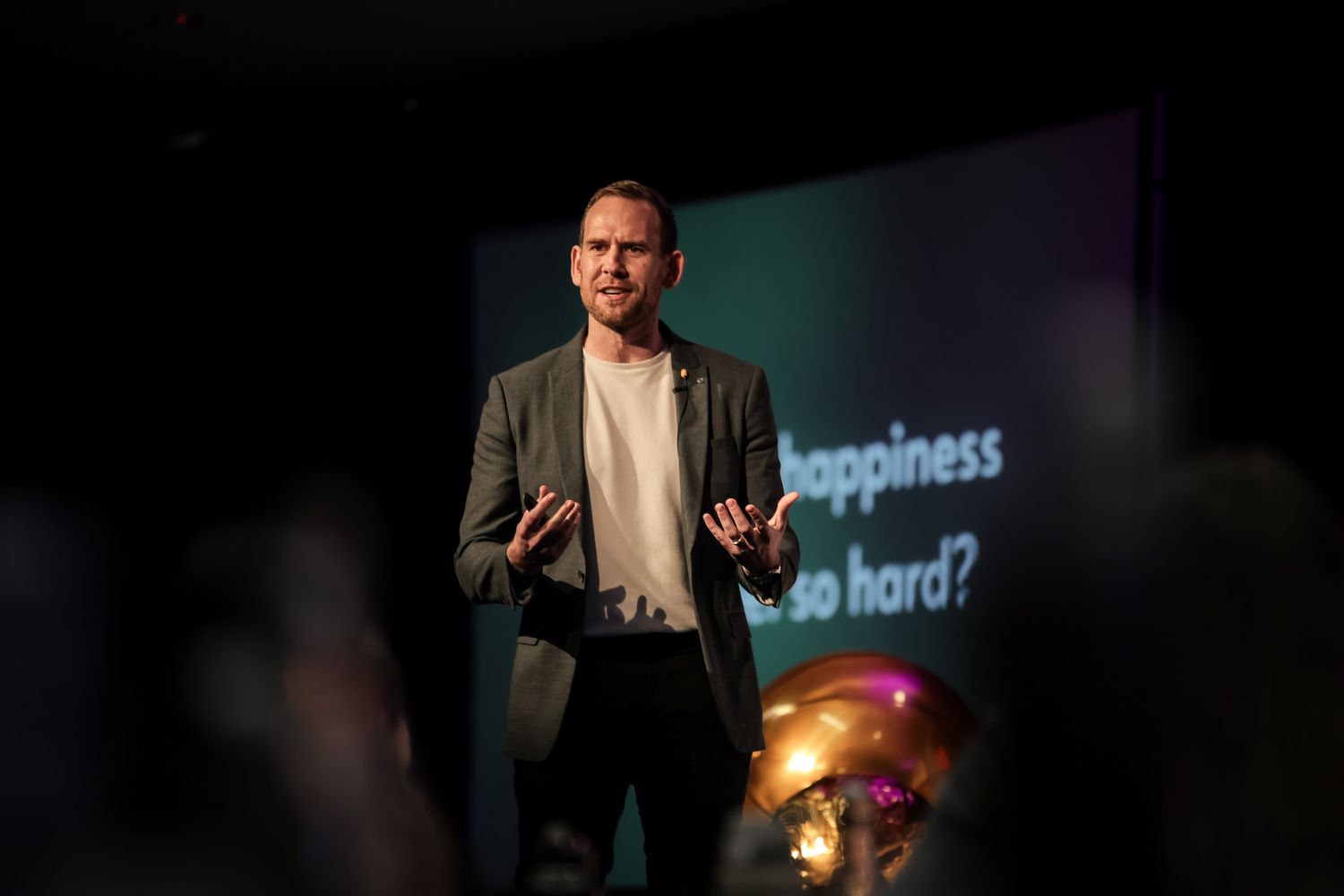 Man in blazer speaking on stage with happiness-themed presentation backdrop in a dimly lit conference setting.