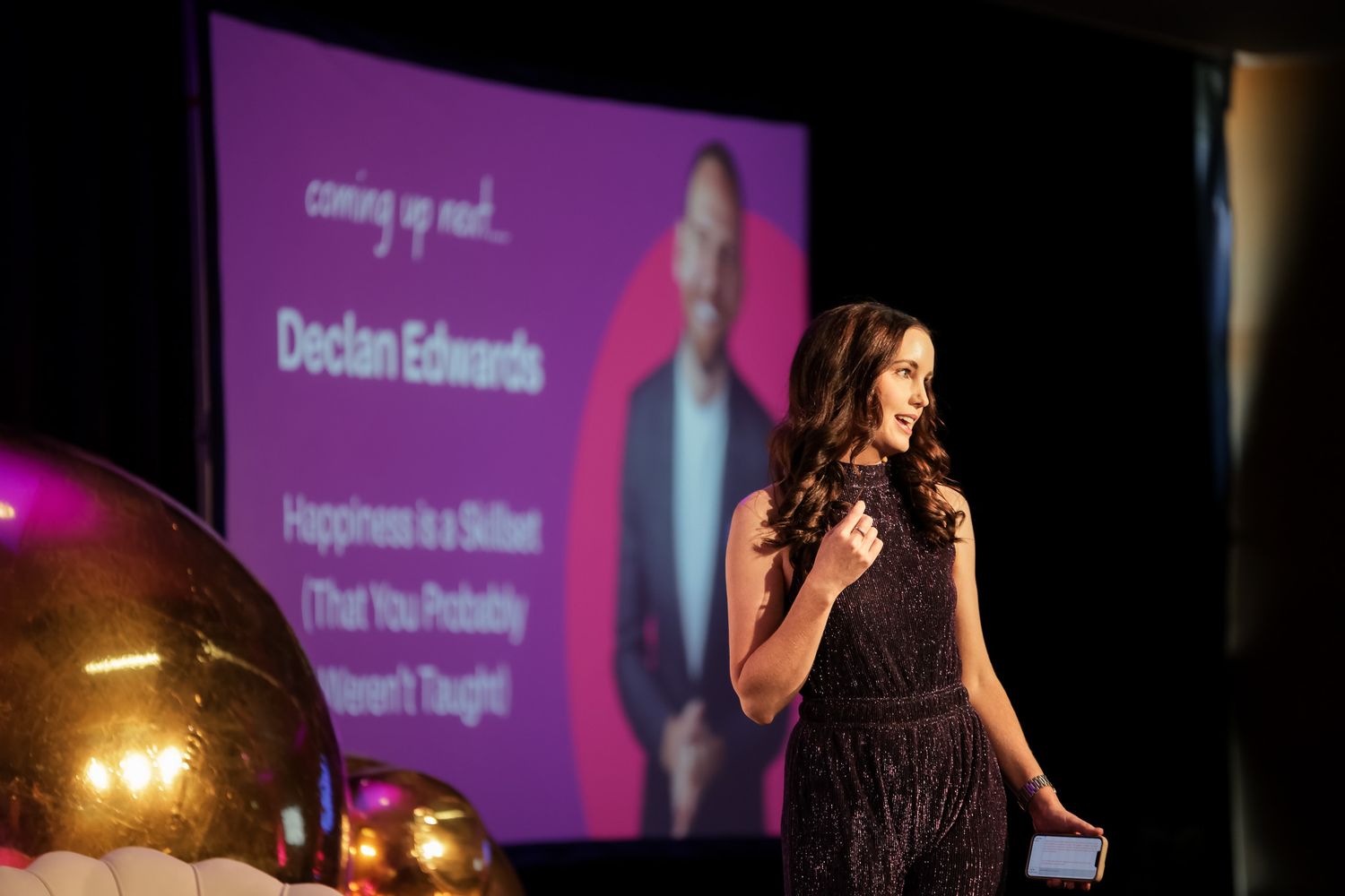 Woman in sparkly dress presenting on stage at an event, with Declan Edwards speaker slide projected behind her.