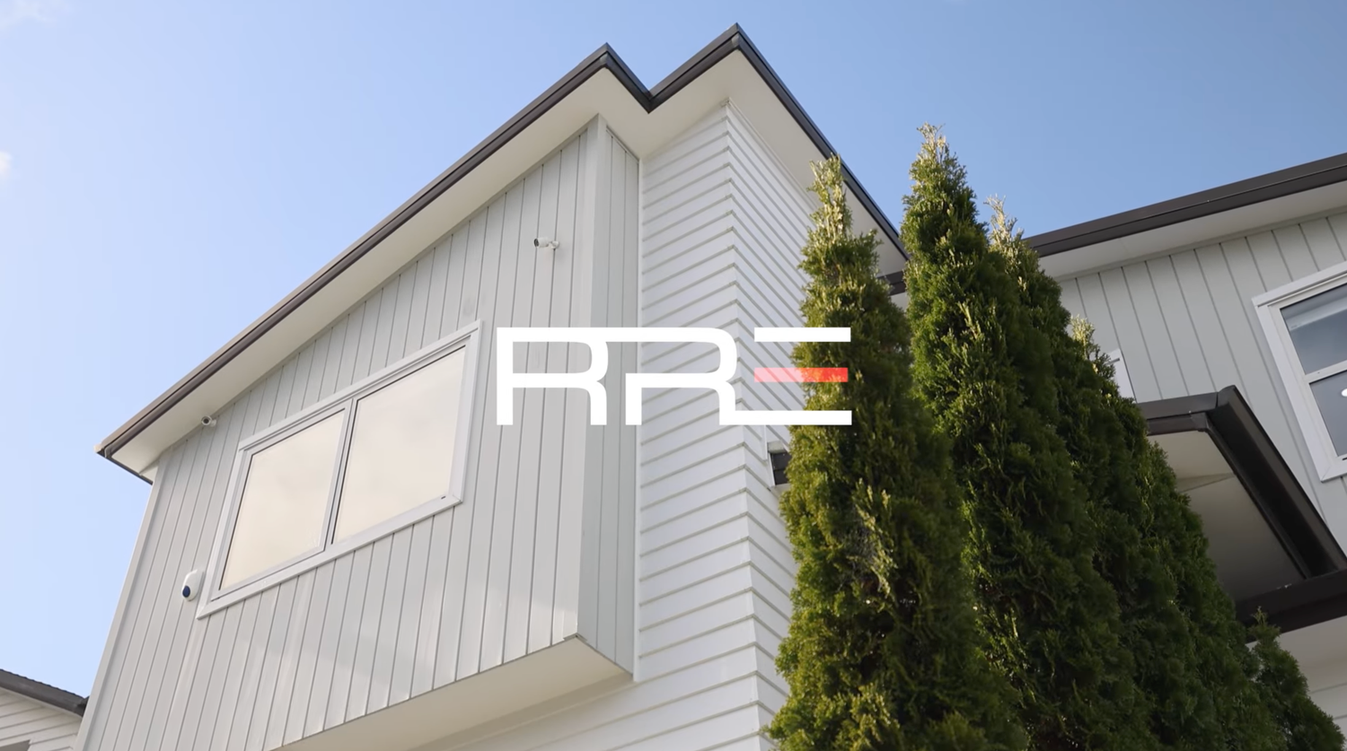 Modern two-story home exterior with white horizontal siding, large windows, and tall evergreen trees under a blue sky.