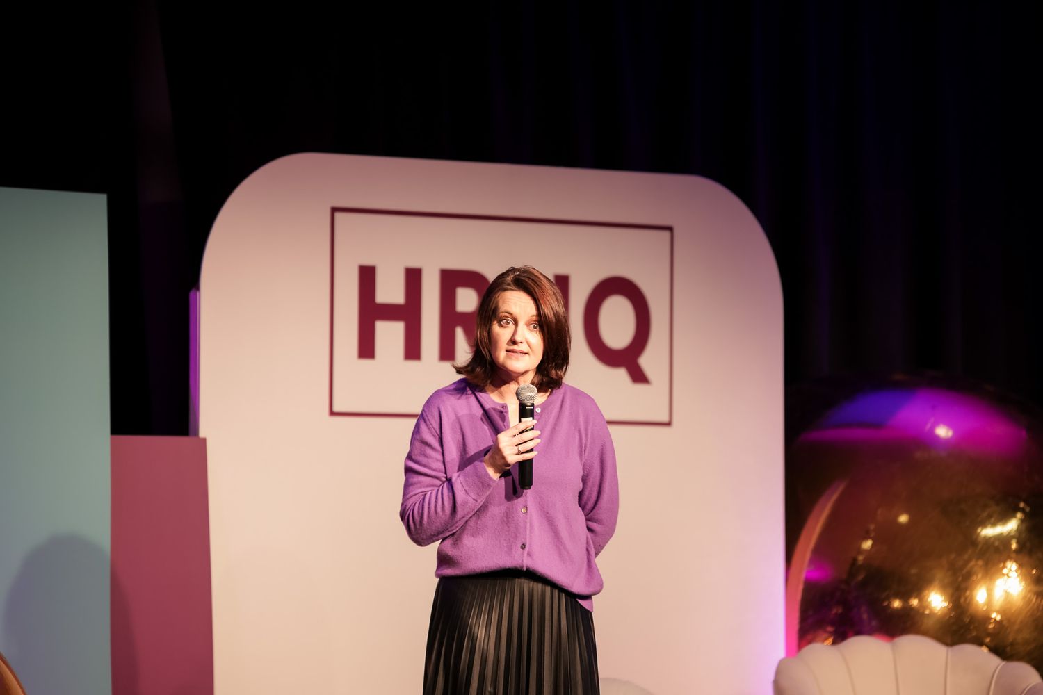 Woman in purple cardigan speaks at HBNQ event, holding microphone on stage with pink backdrop.