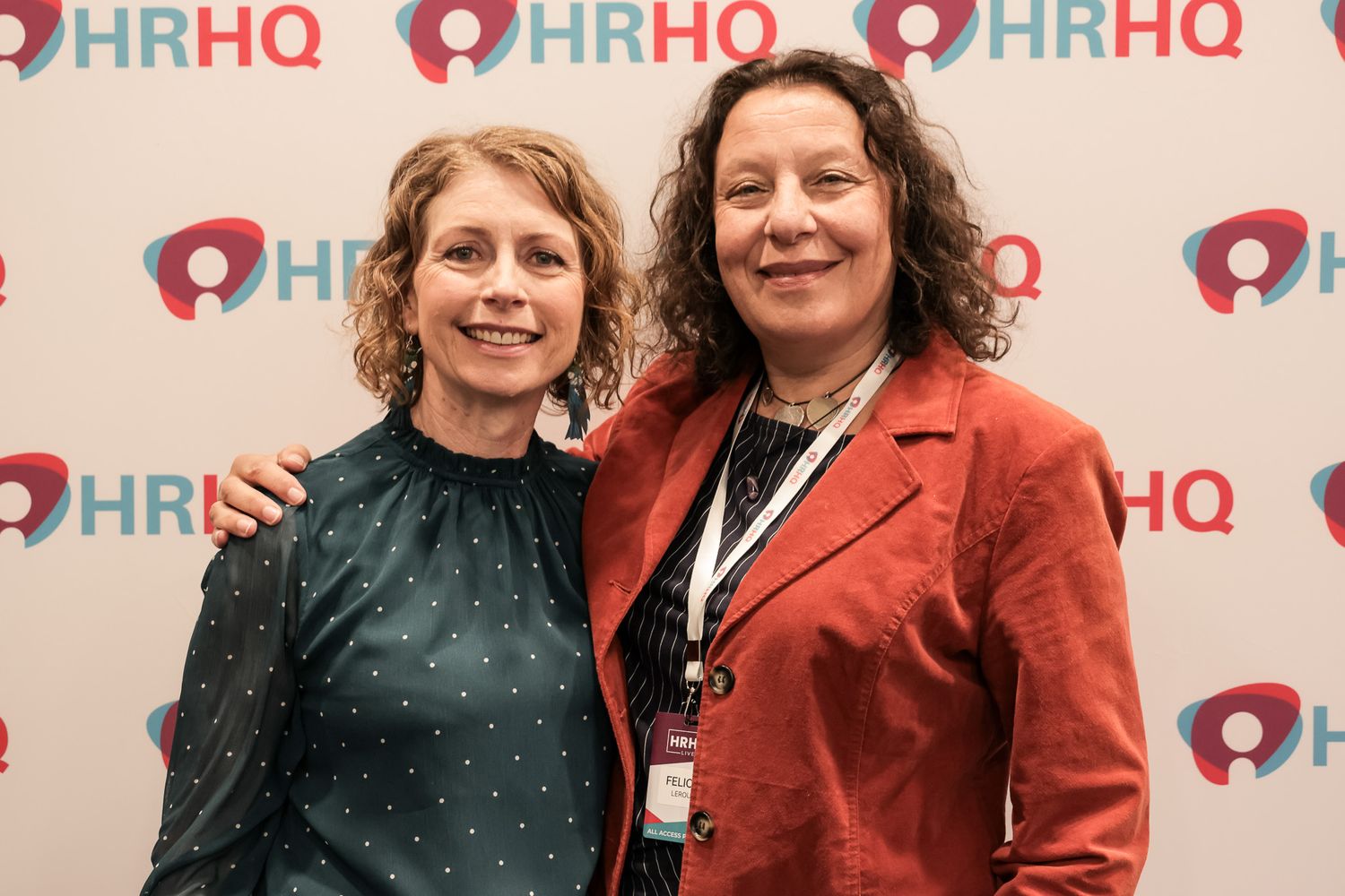Two women smiling together at an HRHQ event, posing in front of a branded step-and-repeat backdrop.