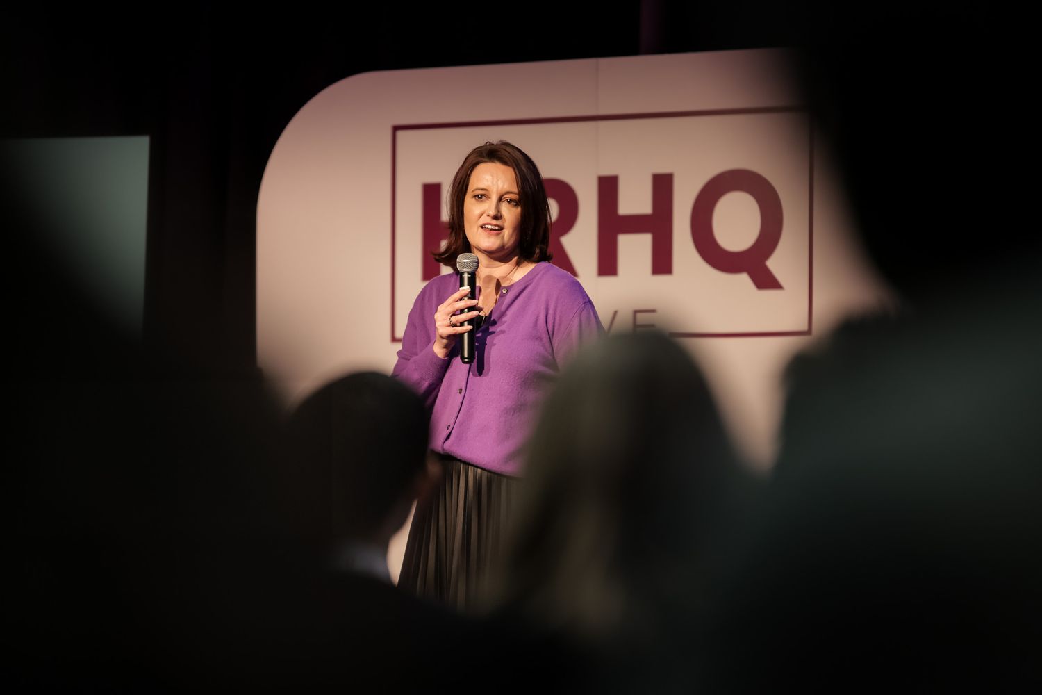Woman in purple blazer speaks into microphone on stage at HPHQ event before seated audience.