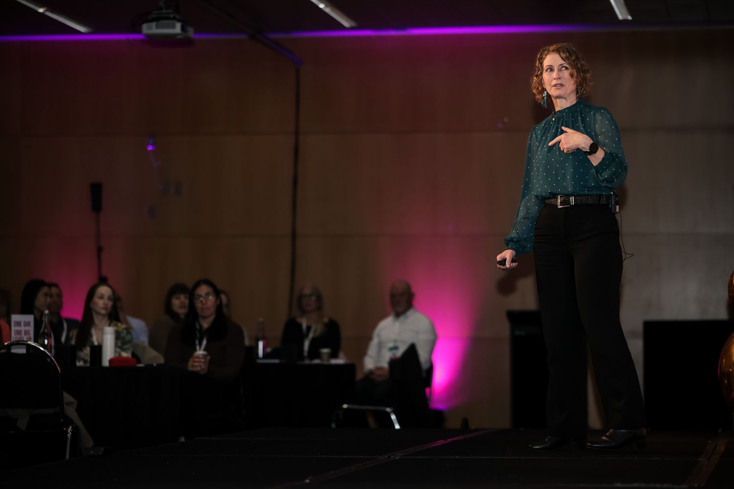 Woman in teal blouse presents on stage at a conference with purple-lit audience seating in background.